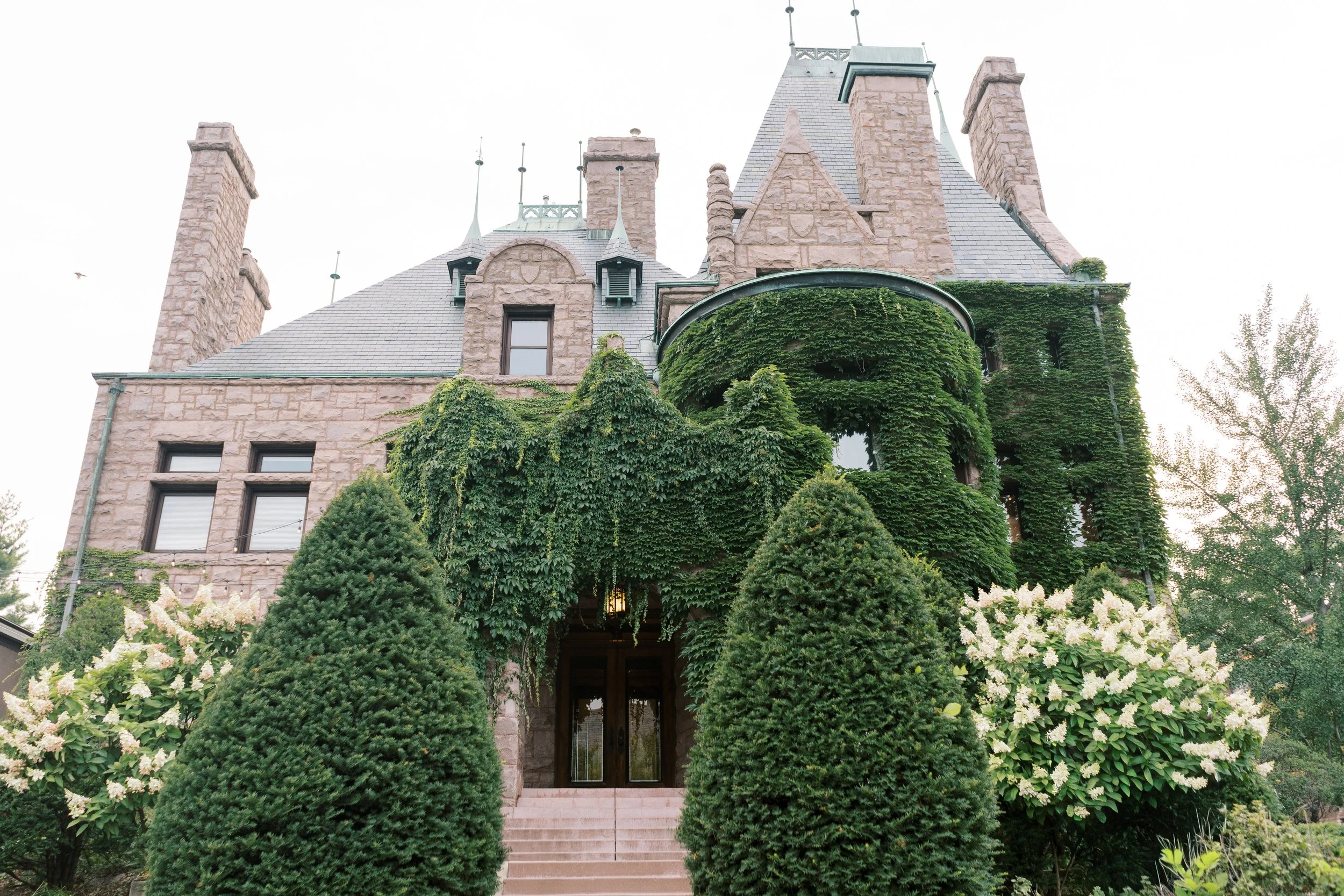 A large stone castle-style house covered in green ivy with neatly trimmed bushes and white flowers in the front yard.