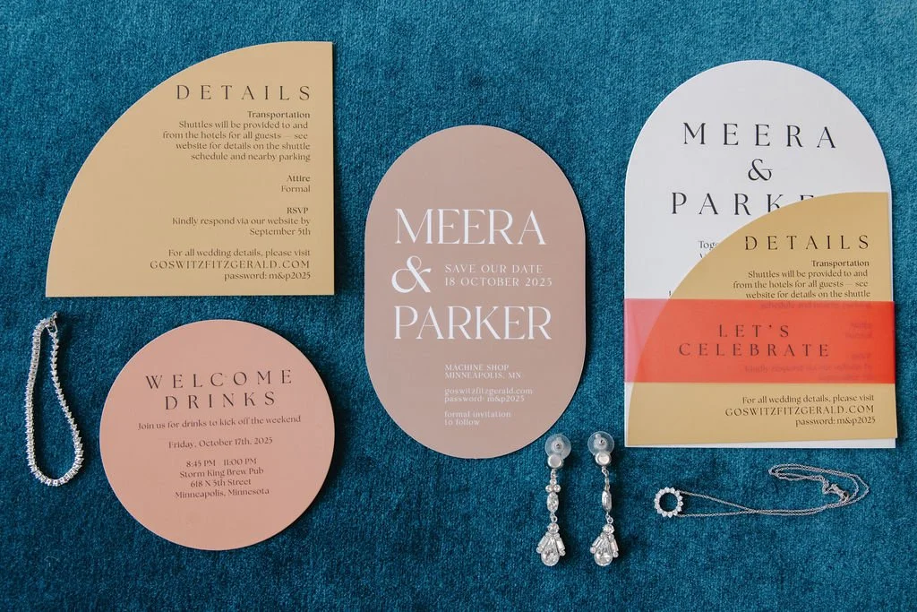 Collection of wedding or event invitation cards including a save-the-date card, a welcome drinks card, a details card, and a celebration card, along with a pair of pearl and rhinestone earrings, and a silver necklace, laid out on a blue surface.