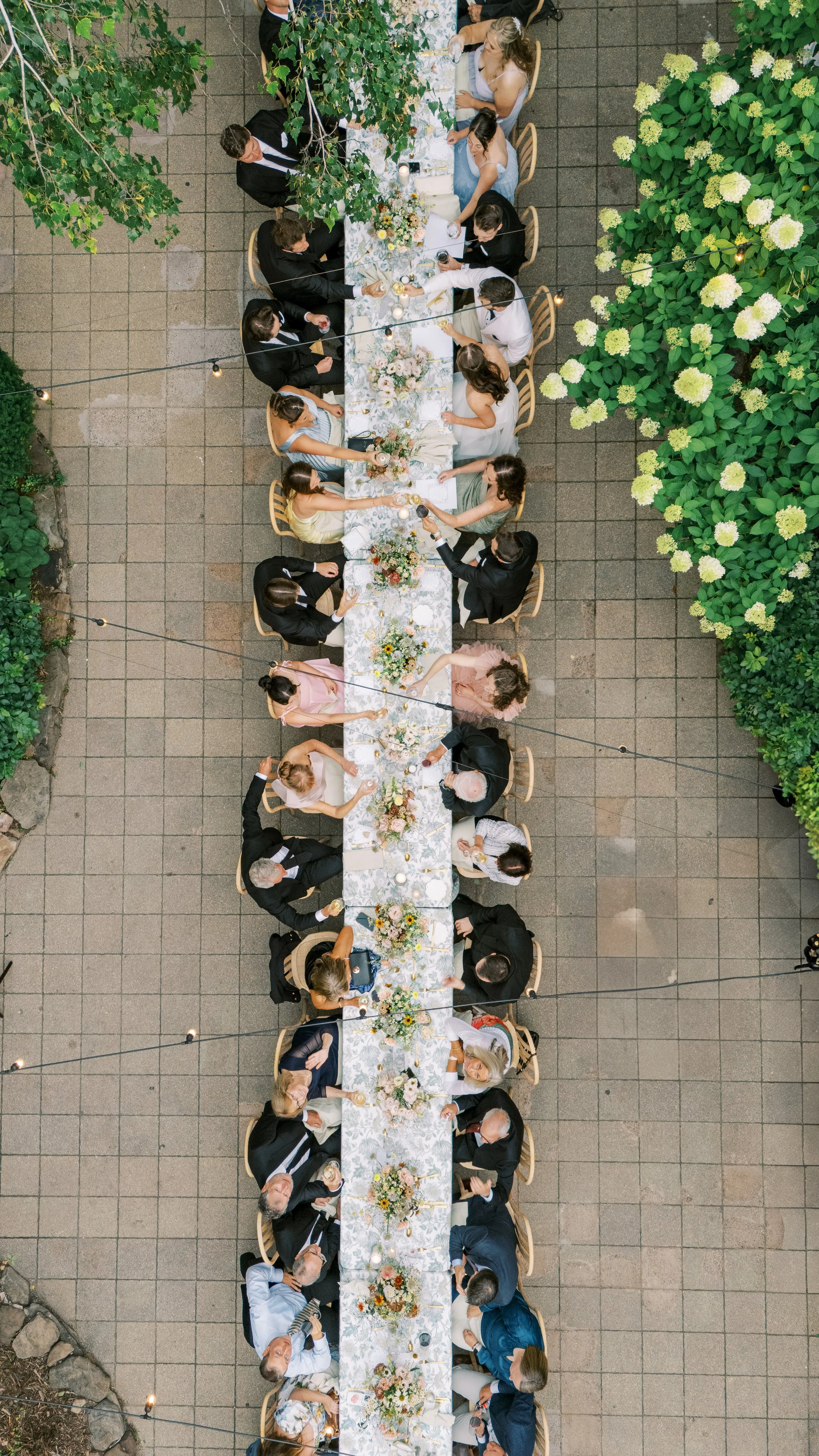 An overhead view of a long outdoor banquet table with floral centerpieces, candles, and guests in formal attire dining and socializing.