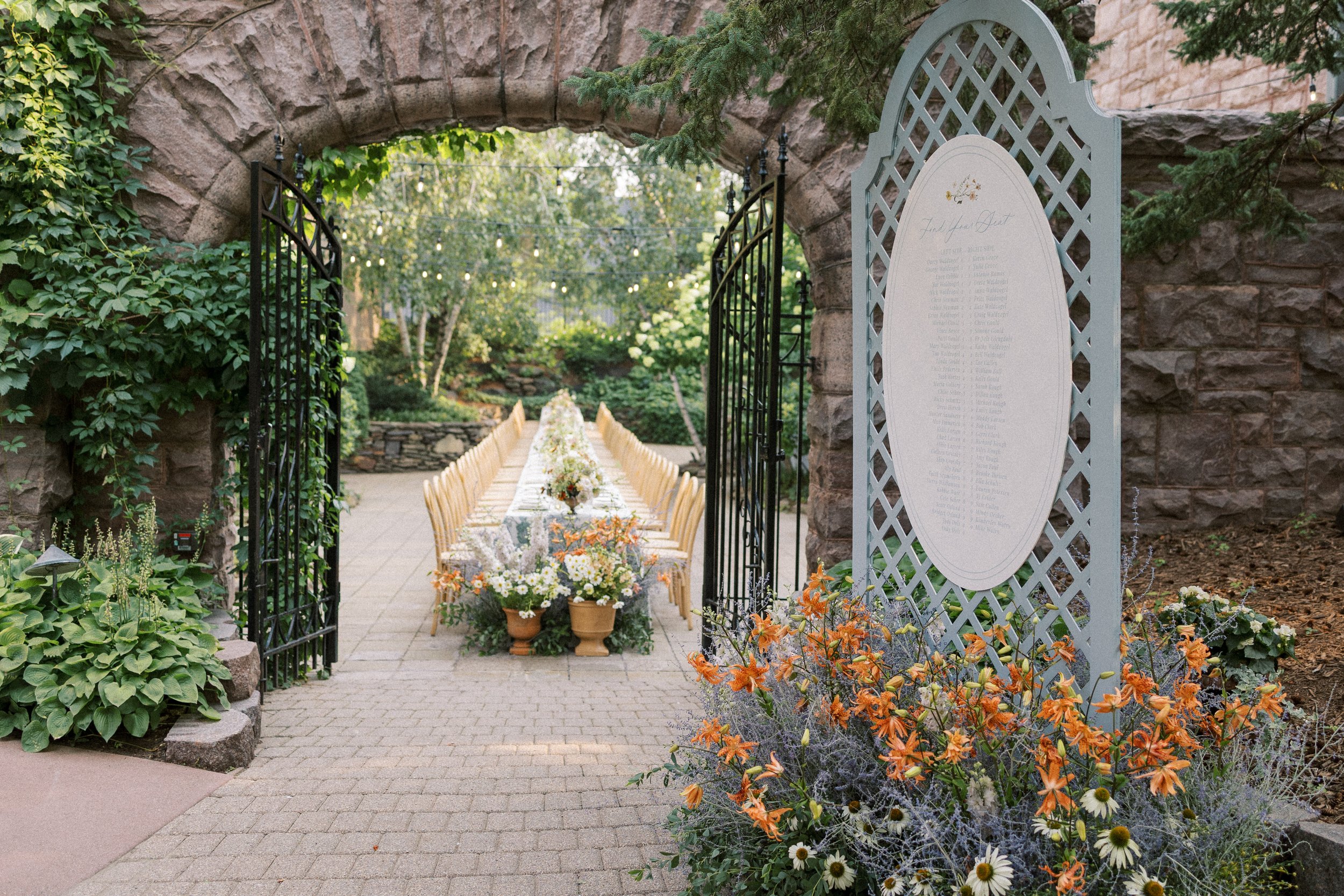 Outdoor wedding reception setup with a long table decorated with flowers, set under a stone archway and a metal gate, surrounded by greenery and string lights, with a seating chart on the right side.