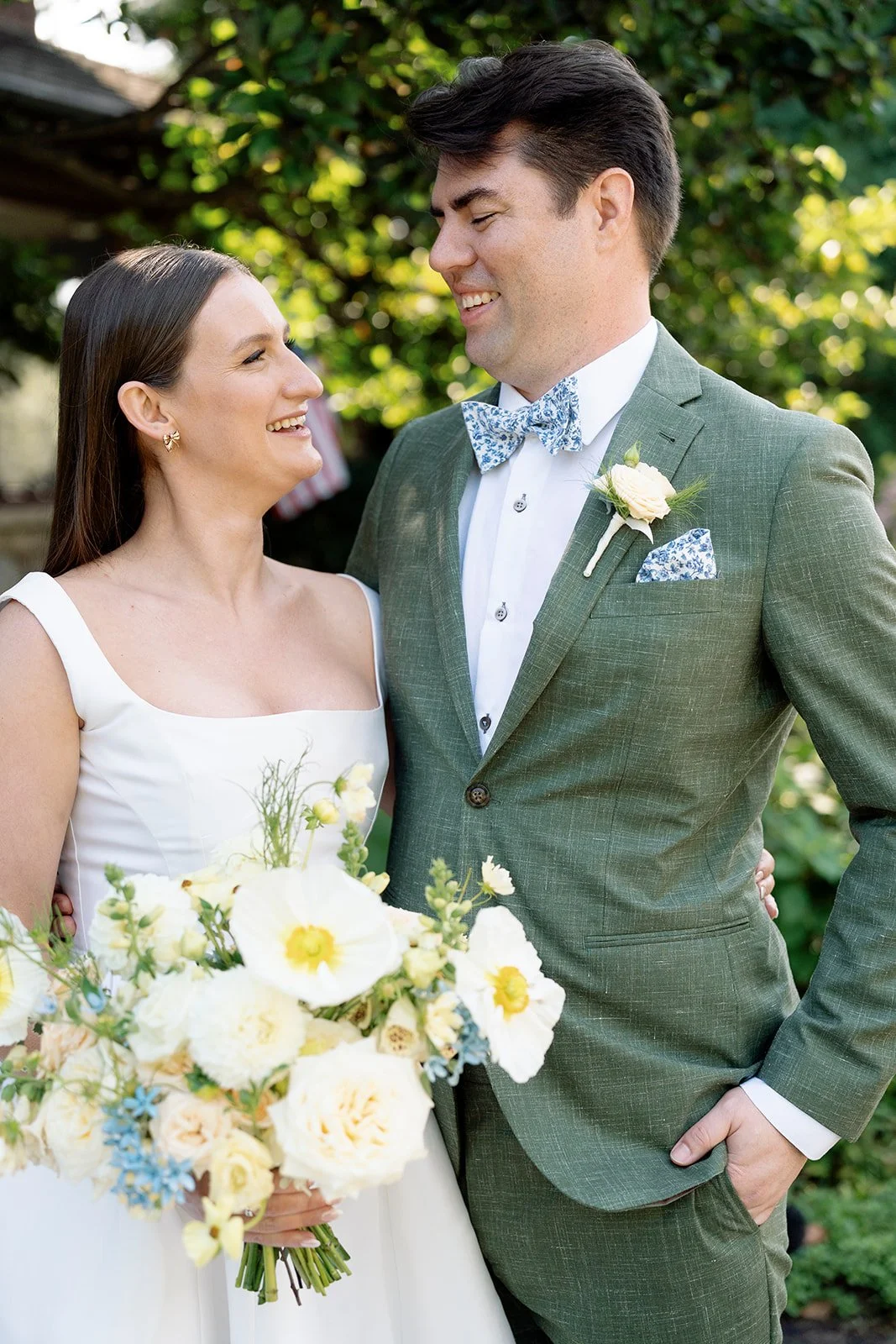 A bride and groom smiling at each other outdoors, with the bride holding a bouquet of white and yellow flowers and the groom wearing a green suit with a floral bow tie and a white boutonnière.
