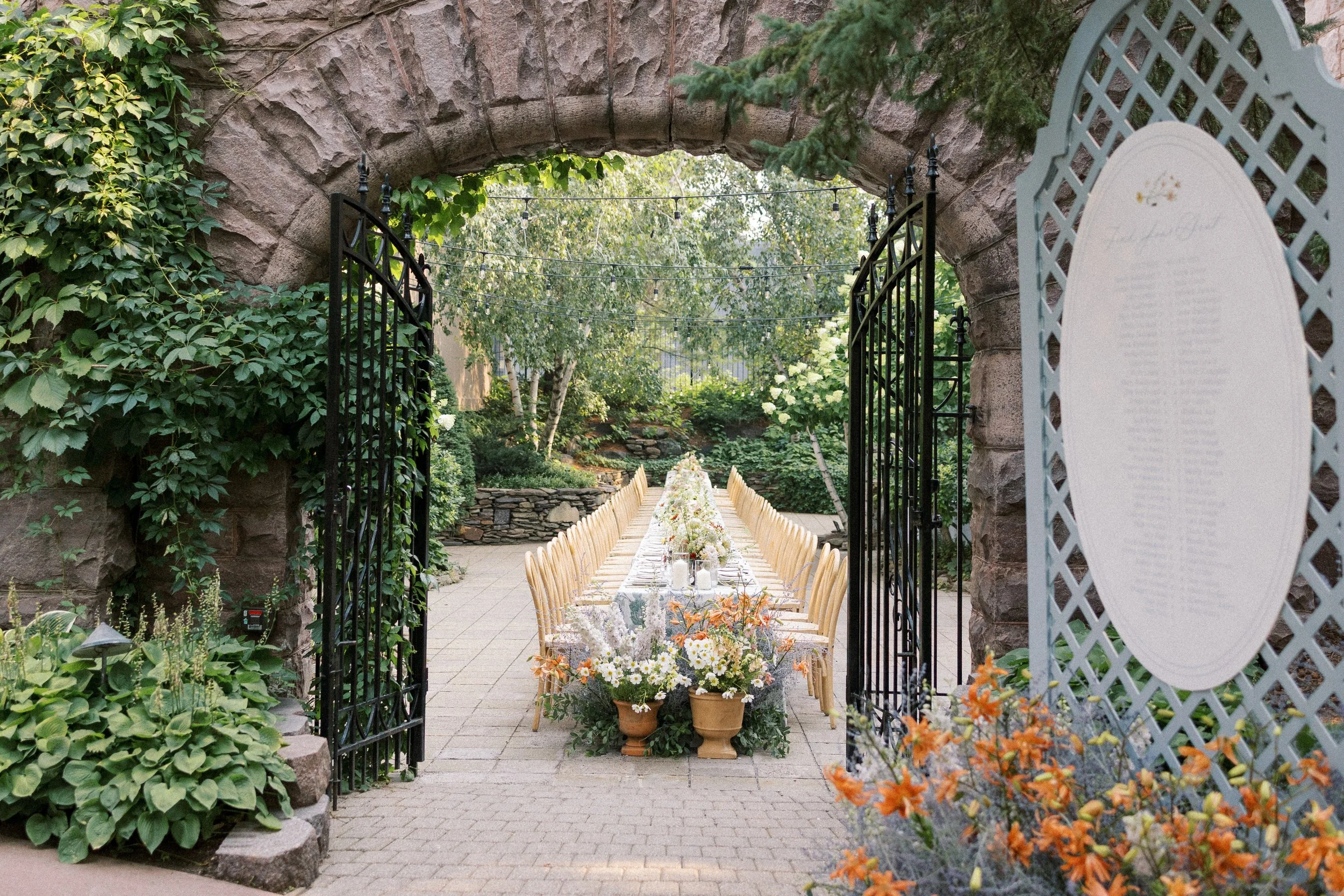 Wedding reception setup in a garden with a long table decorated with flowers, surrounded by wooden chairs, viewed through a decorative gate under a stone archway.