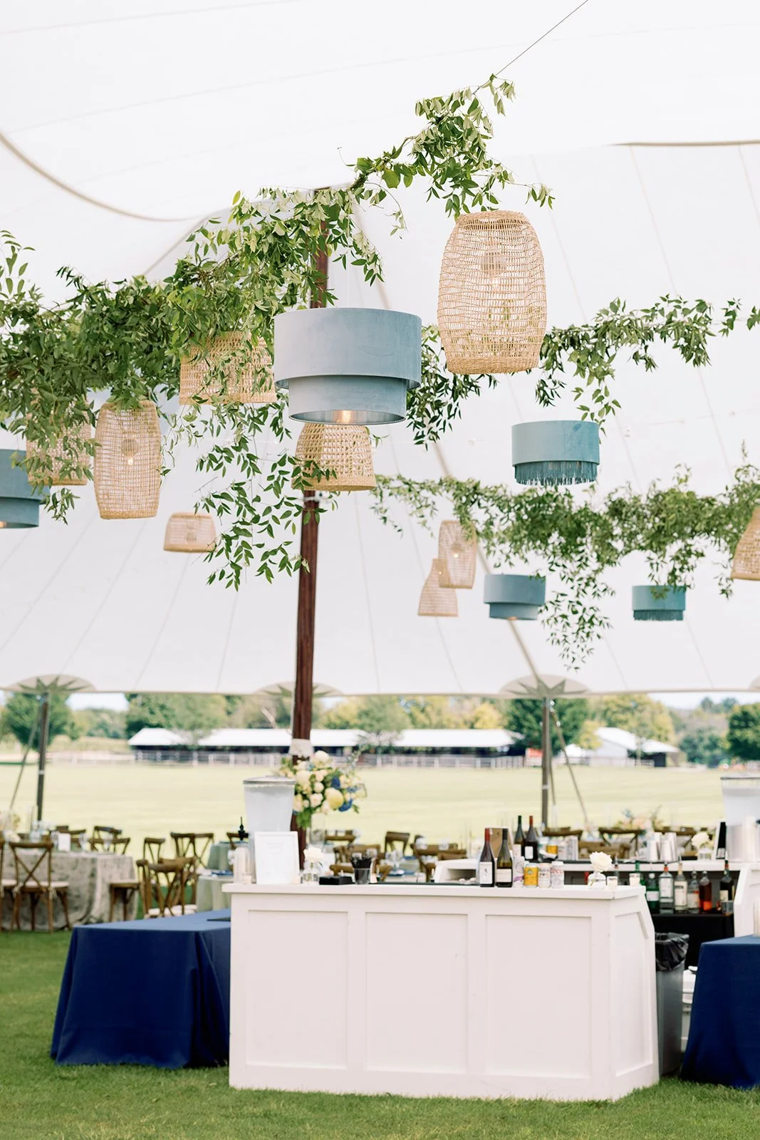 Outdoor event setup with hanging lanterns and greenery under a large tent, with tables and chairs in the background.