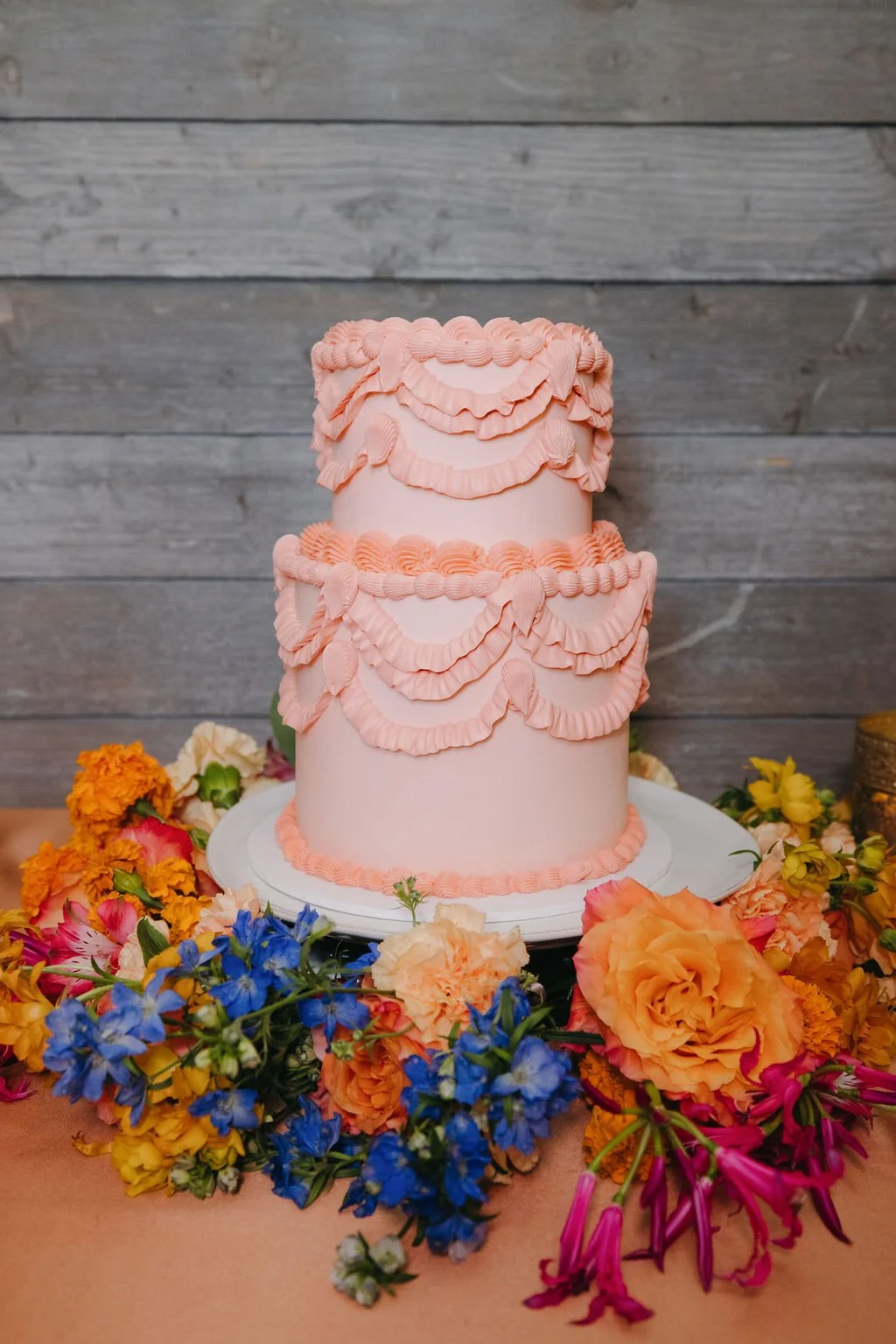 A three-tier pink cake decorated with ruffled icing, surrounded by colorful flowers including orange, yellow, pink, and blue blooms, on a table against a wooden background.