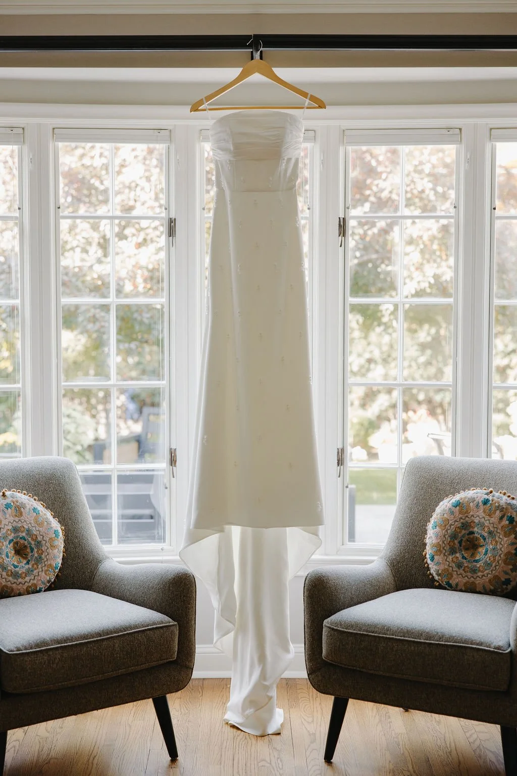 A white wedding dress hanging from a wooden hanger on a black curtain rod, in front of large window with multiple panes.