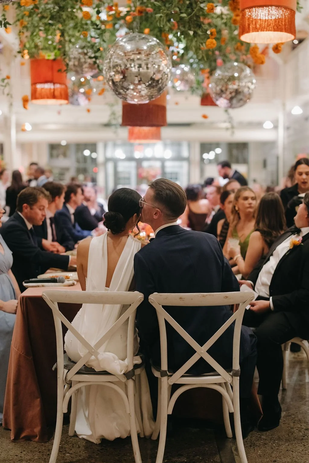 A bride and groom share a kiss at the center of a wedding reception with guests seated around them at long tables, decorated with floral arrangements and hanging decorations.