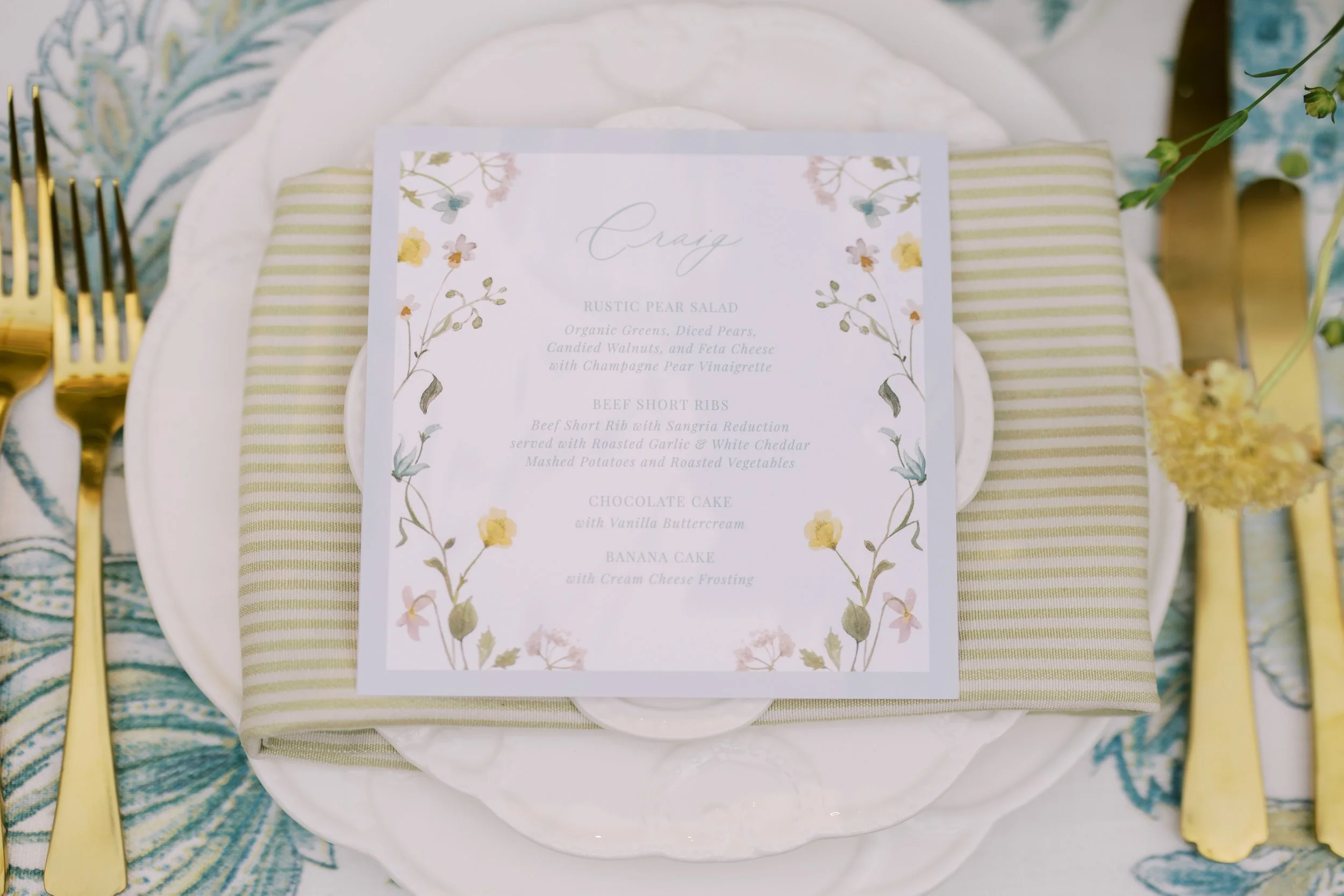 Wedding place setting with a menu card, gold utensils, a floral tablecloth, and a small flower arrangement.