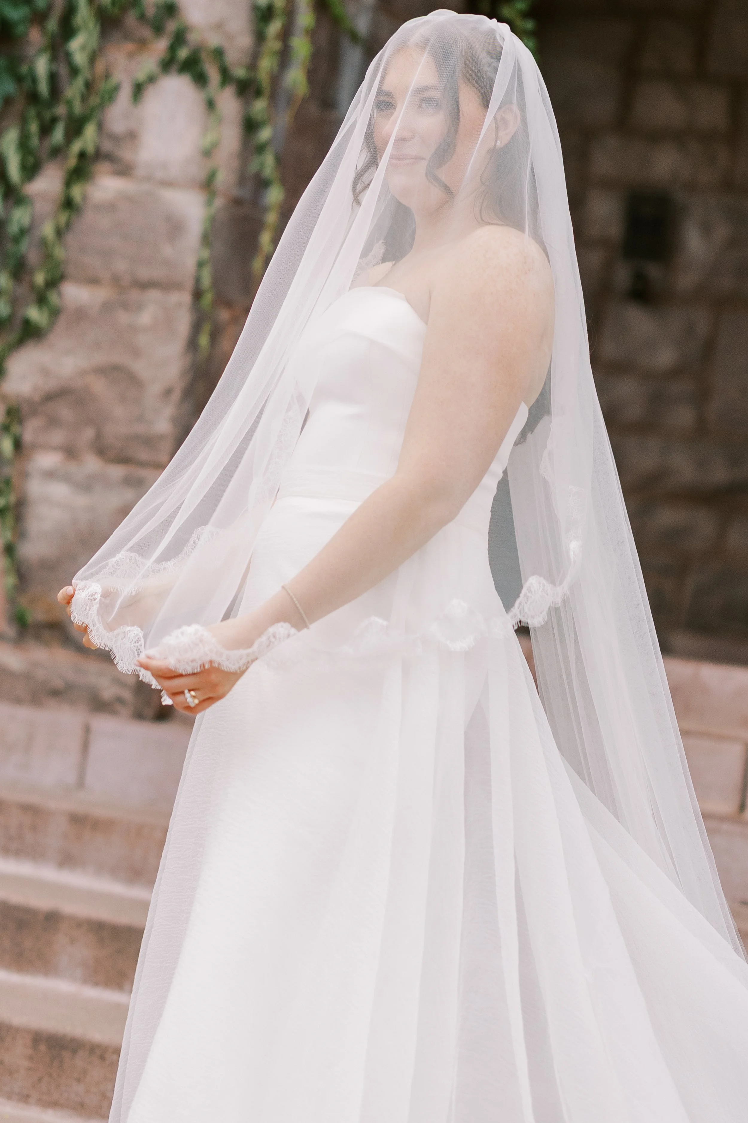 A woman in a white wedding dress and veil standing outdoors in front of a stone wall with steps, holding the veil with both hands.