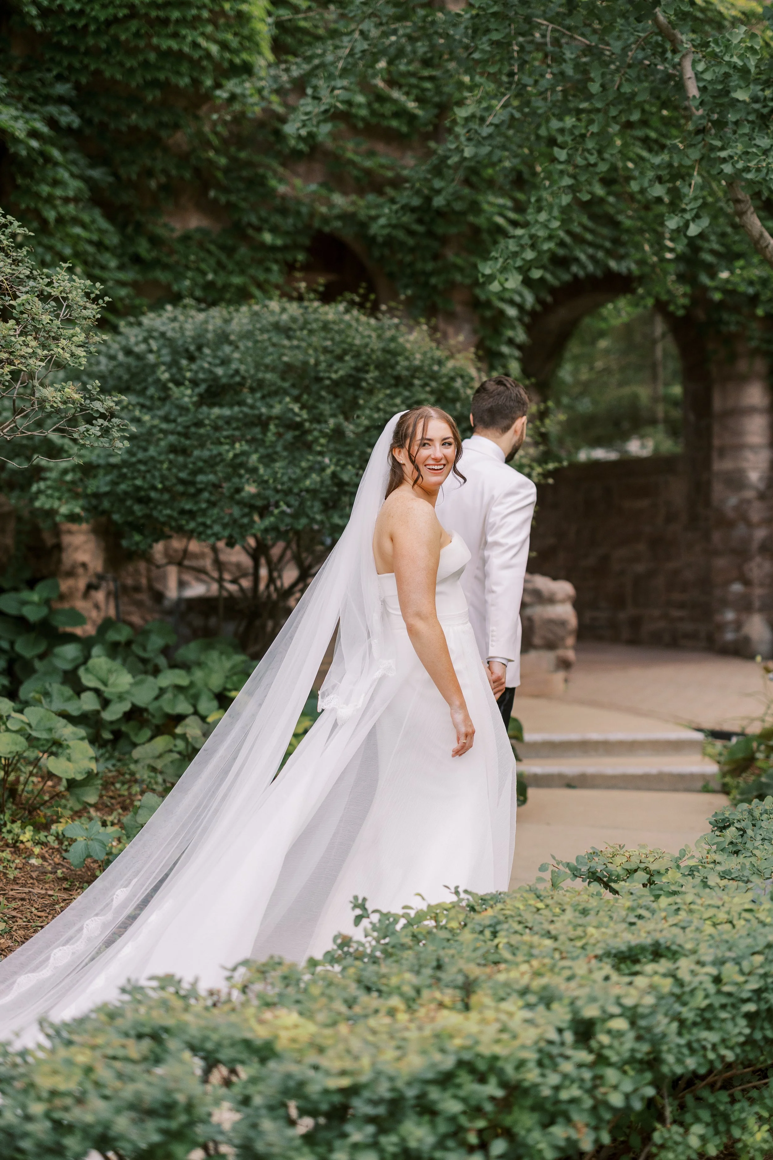 Bride in a white wedding dress smiling, looking back at the camera, with a groom standing beside her, outdoors with greenery and stone arch in the background.