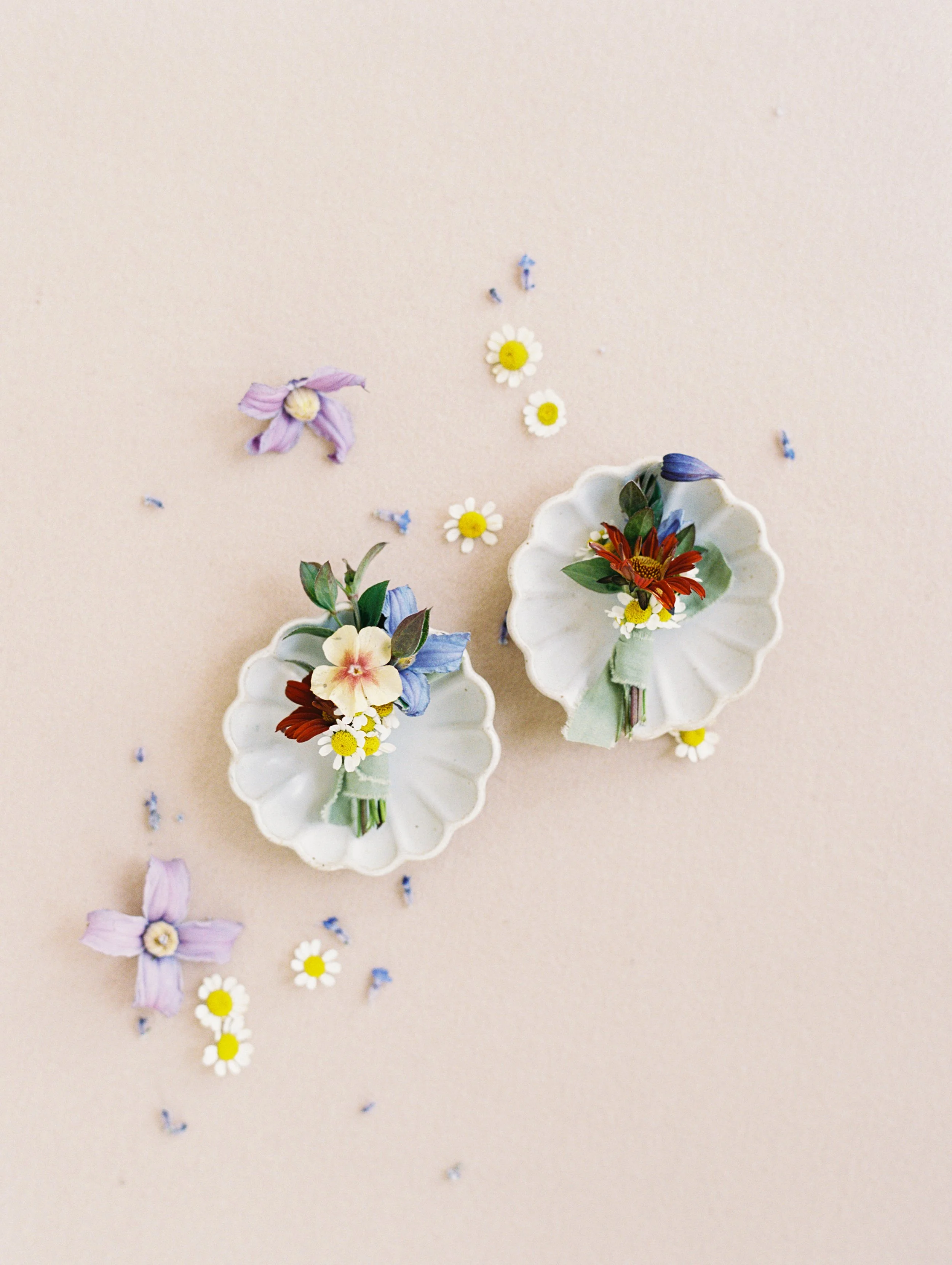 Two white scalloped ceramic dishes with small bouquets of flowers, surrounded by scattered small flowers and petals on a light pink background.