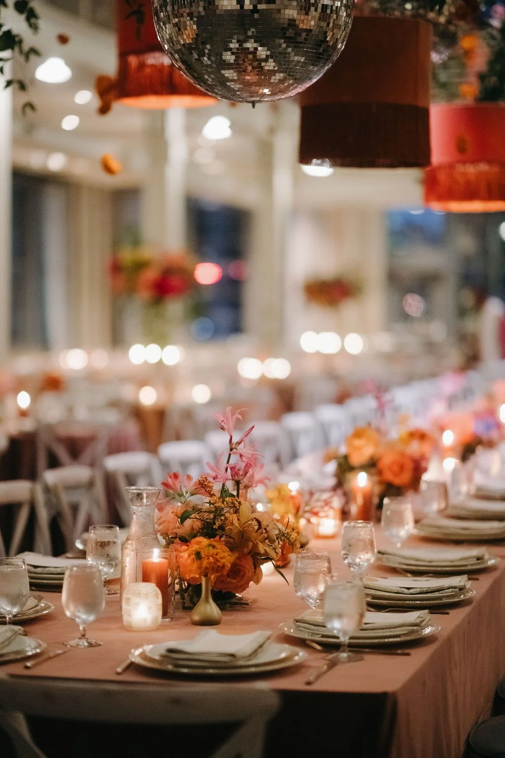 Elegant banquet table decorated with orange and pink flowers, candles, and tableware, under hanging disco ball and colored lanterns in a warmly lit venue.