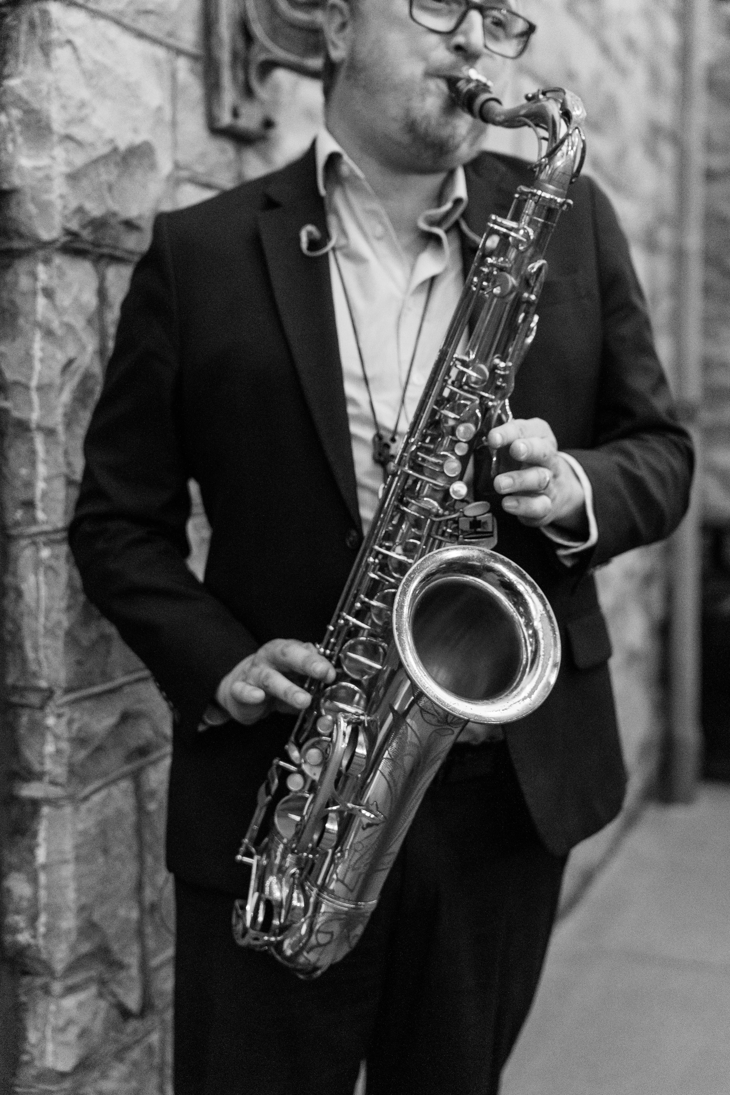Man in a suit playing a saxophone against a brick wall.