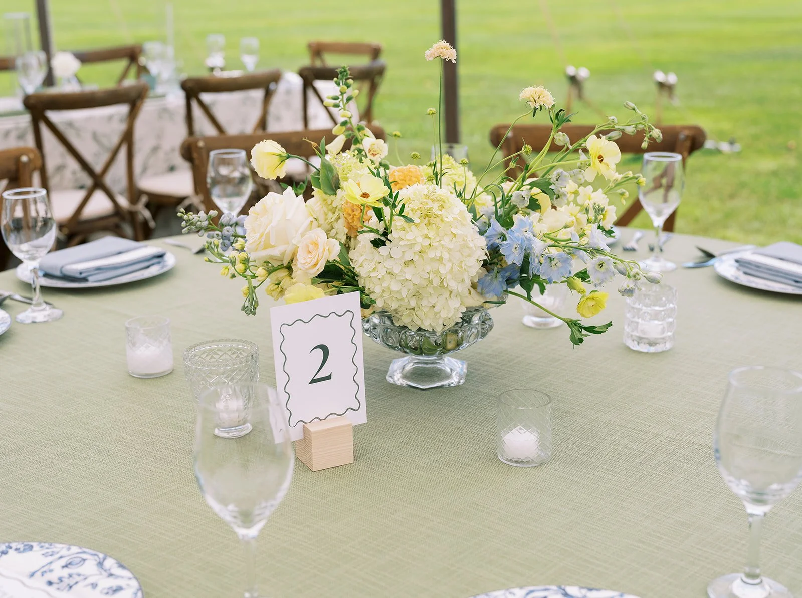 A dining table decorated with a large white floral centerpiece, set with glassware, plates, and candles, at an outdoor event.