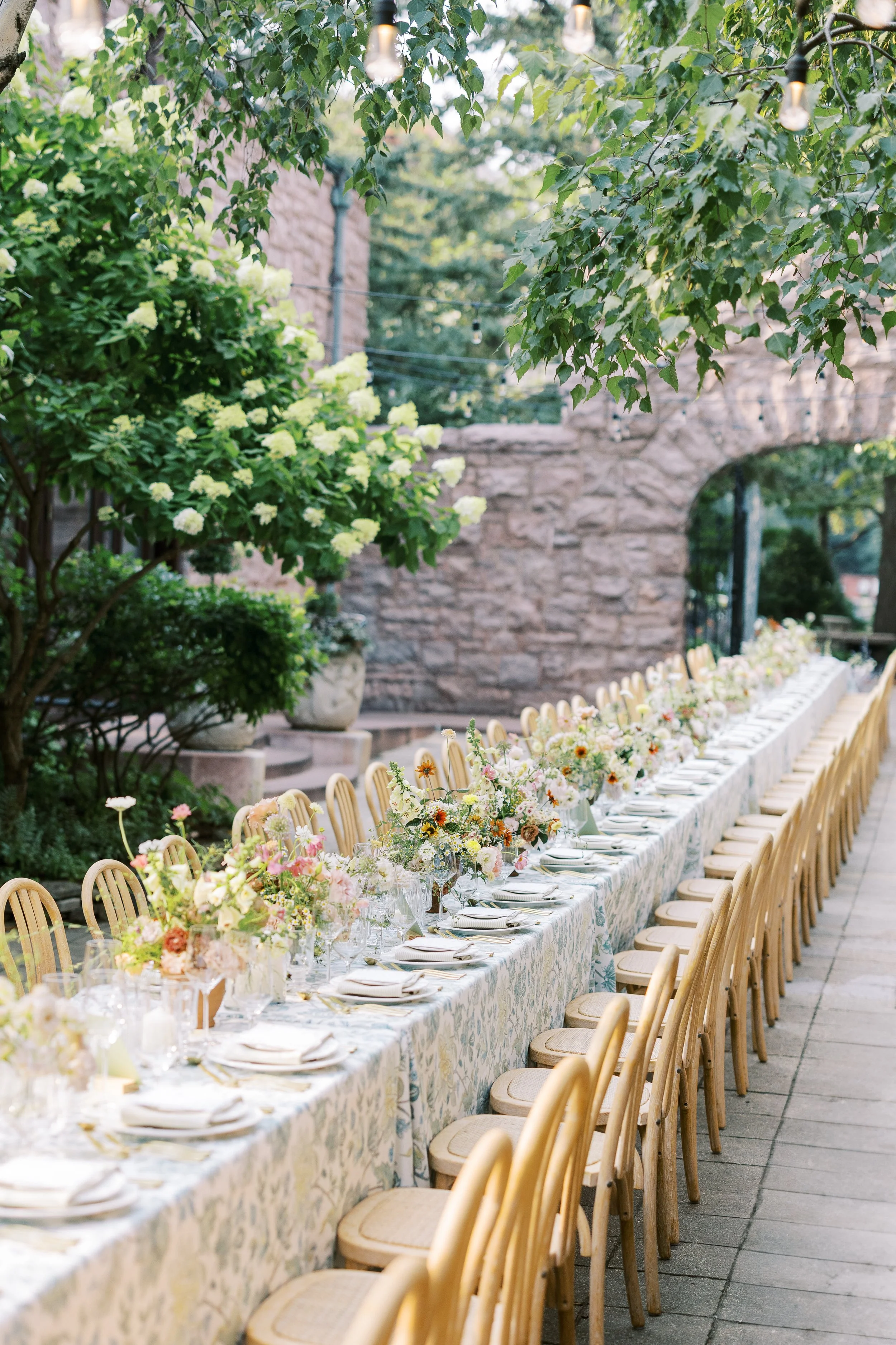 Long outdoor dining table decorated with floral centerpieces, set with plates, glasses, and napkins, surrounded by wooden chairs, in a garden with green trees and string lights.
