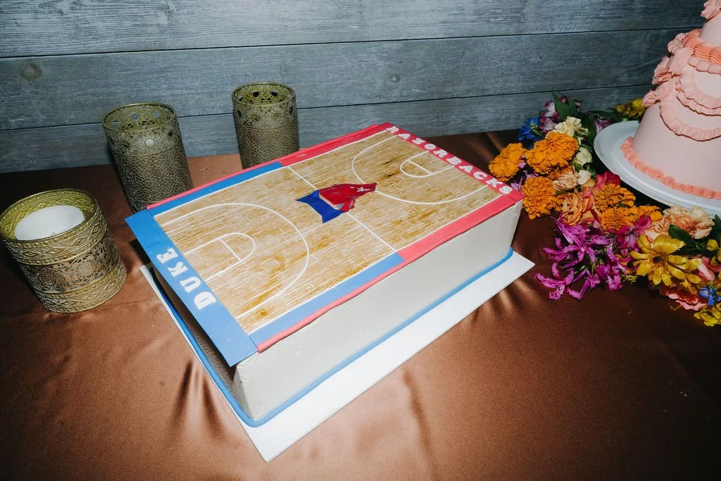 Cake decorated as a basketball court for a basketball-themed celebration, with the words 'DUE' and 'ON' on the sides, placed on a table with candle holders and surrounded by colorful flowers.