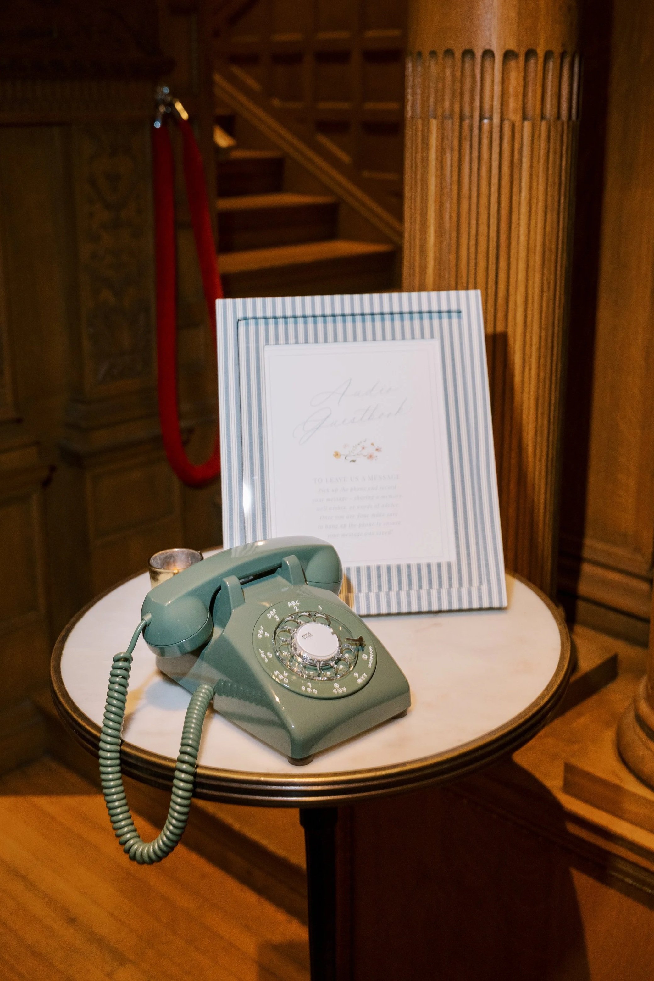 A vintage mint green rotary telephone on a small round table, with a framed sign behind it, in a wooden-paneled room.