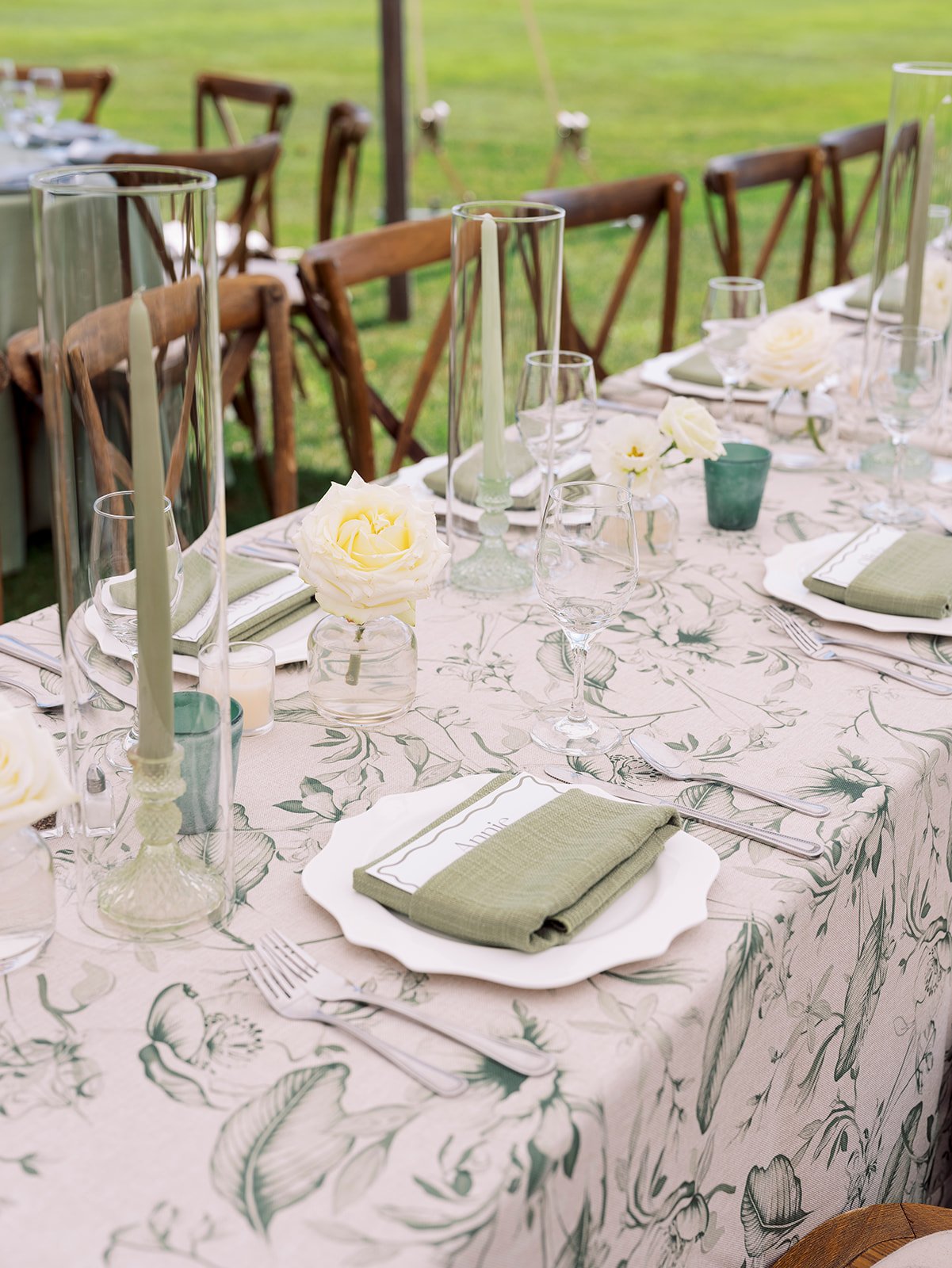 Elegant outdoor table setting with white floral-patterned tablecloth, green napkins on white plates, tall glass candle holders, small floral arrangements, various wine glasses, and wooden chairs surrounding the table on a grassy lawn.