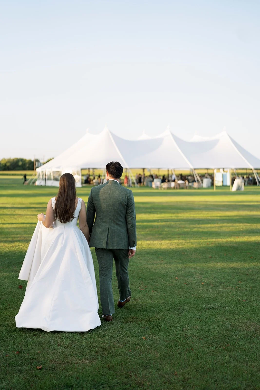 A bride and groom walking away on a grassy field towards a large white event tent in the distance.
