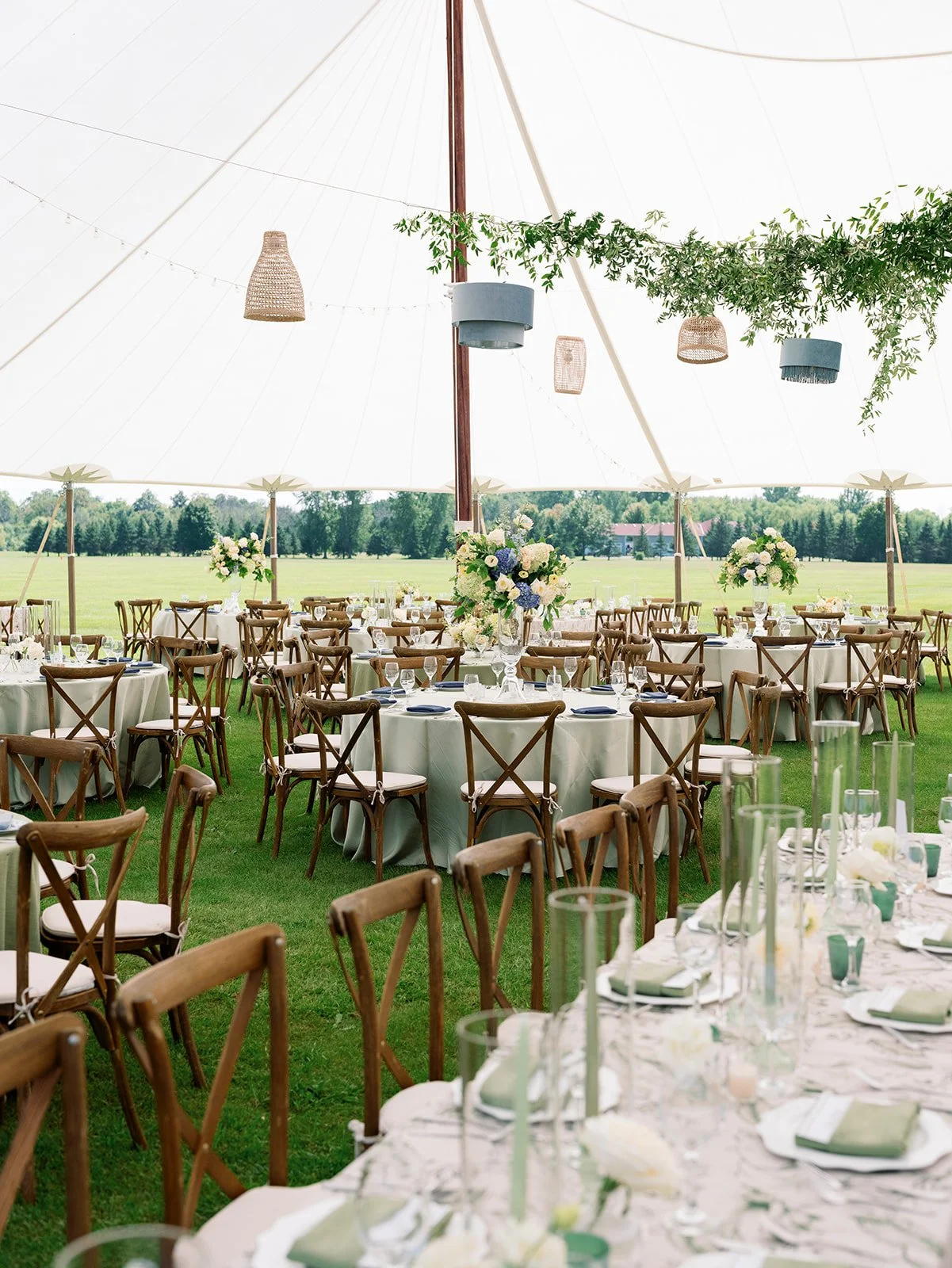 Wedding reception setup with round tables, wooden chairs, floral centerpieces, and a large outdoor tent on a grassy field.