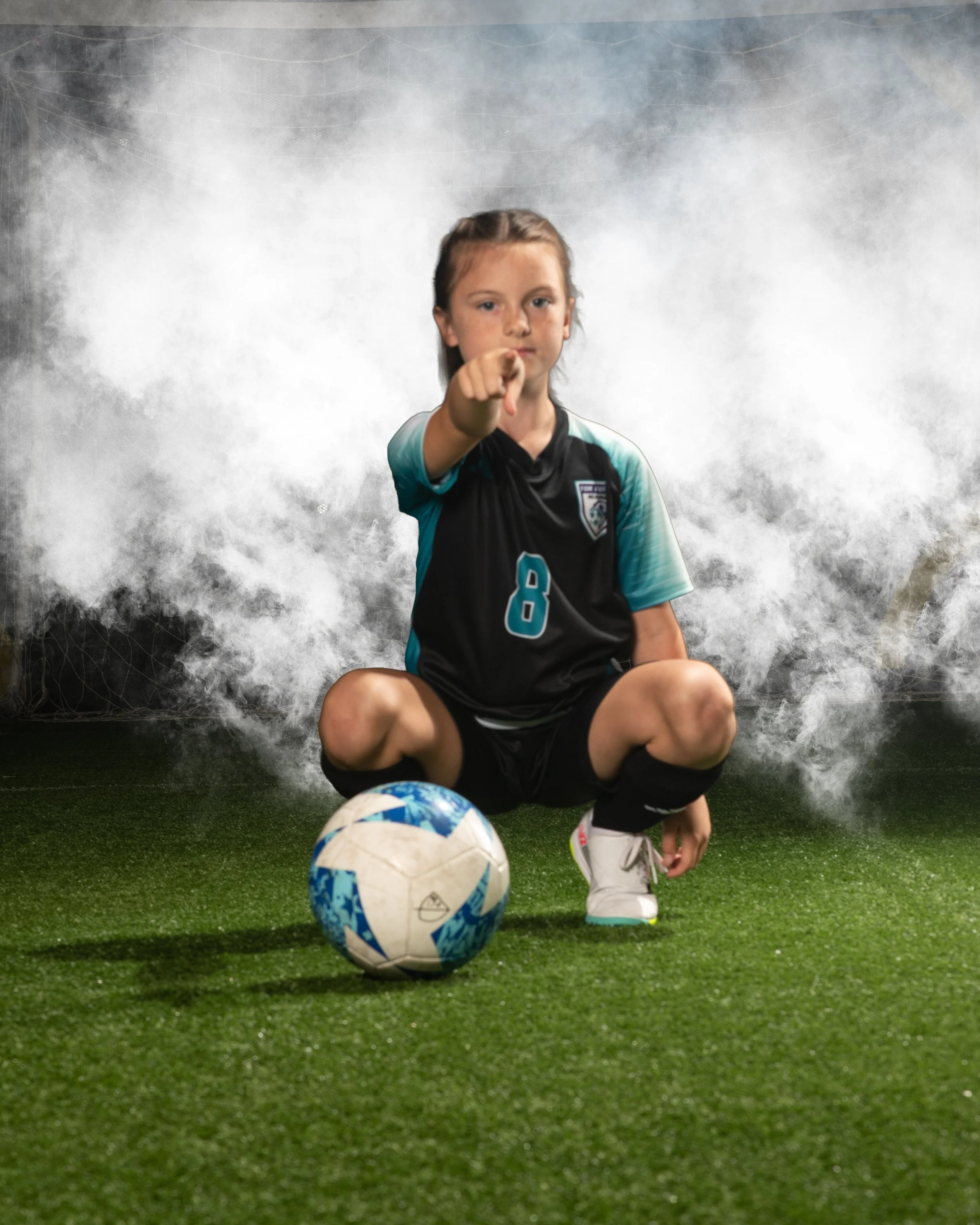 Young girl in a soccer uniform squatting with a soccer ball, pointing at the camera, with a smoky background.