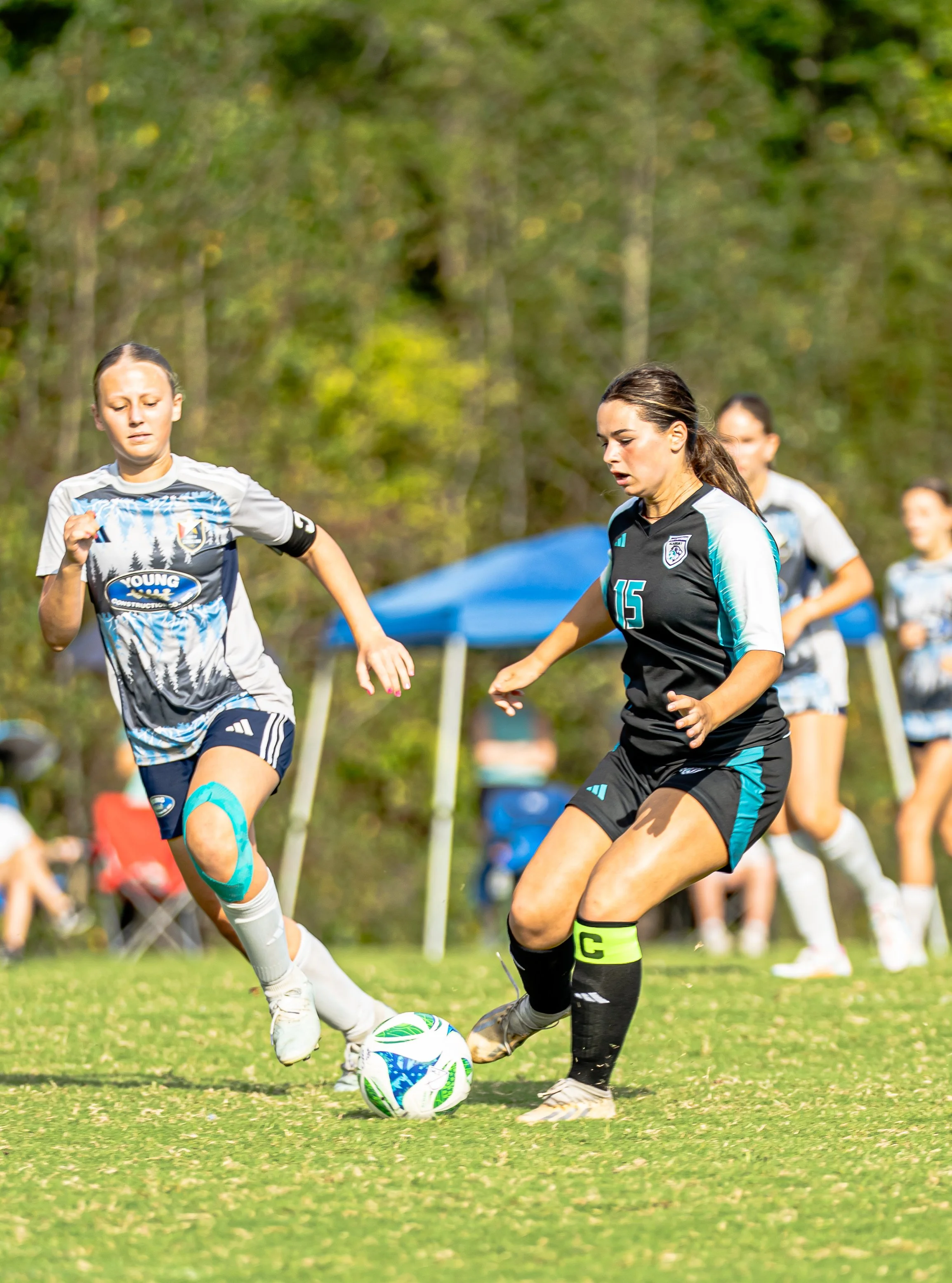 Two female soccer players competing for the ball during a match on a grassy field with trees in the background.
