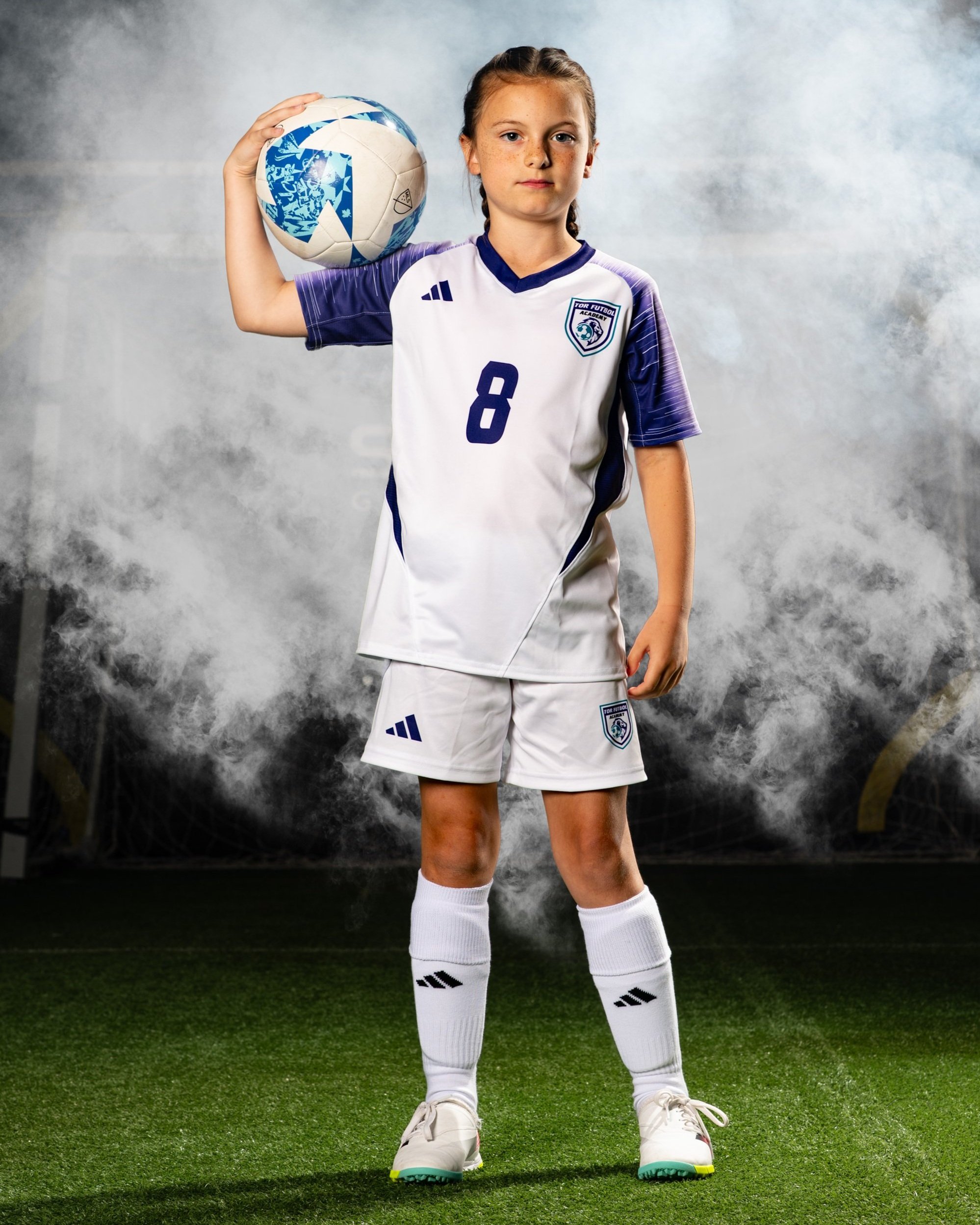 A young girl in a soccer uniform holding a soccer ball on her shoulder, standing on a soccer field with a cloudy background.