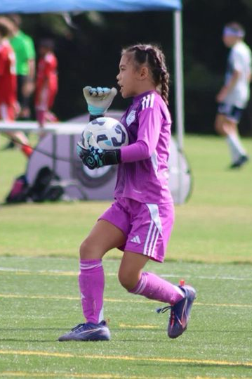 Young girl in a purple soccer uniform holding a soccer ball, running on a soccer field during a game.