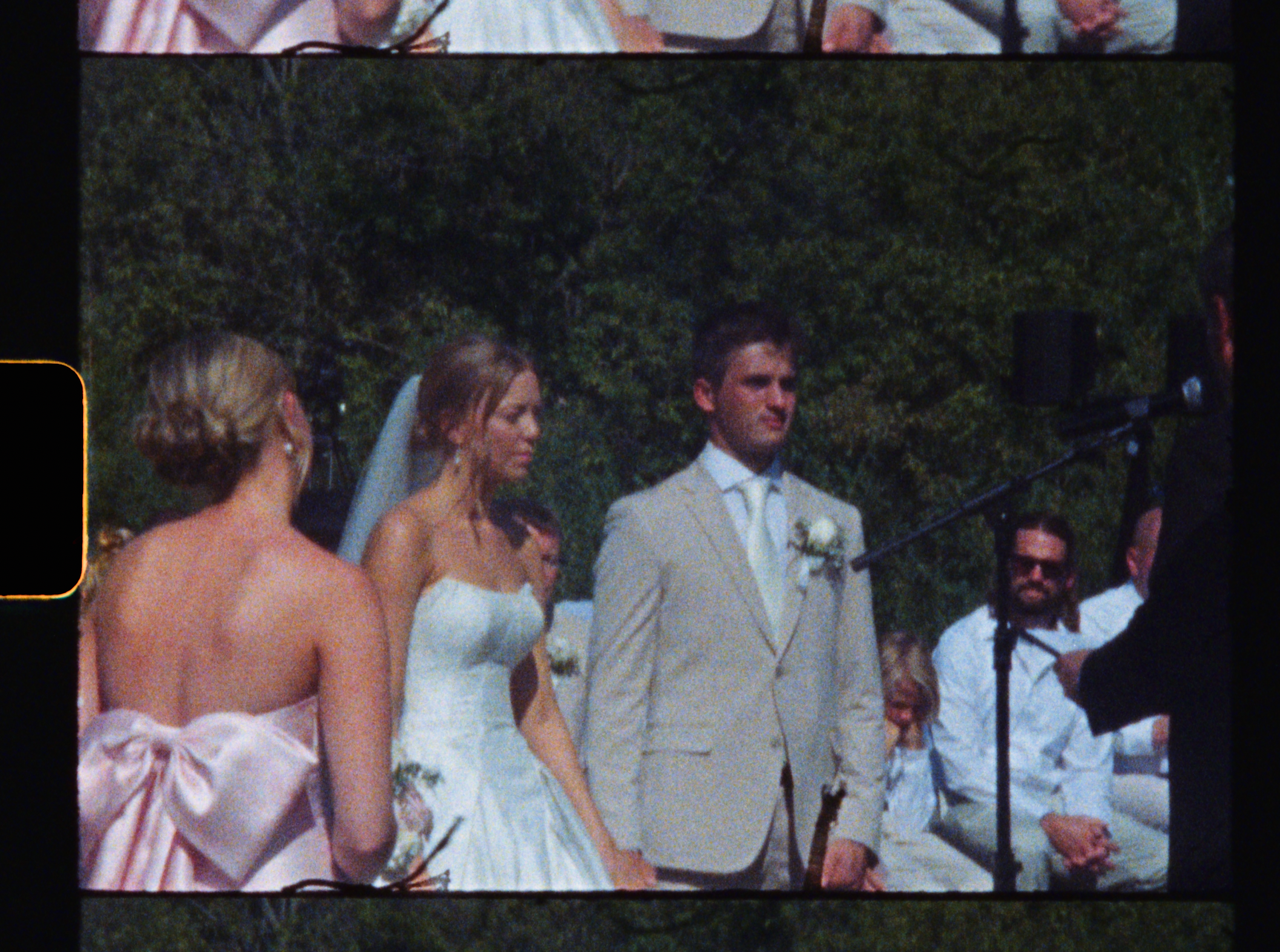 A wedding ceremony outdoors with a bride, groom, and guests standing, with a microphone and speaker visible, and greenery in the background.