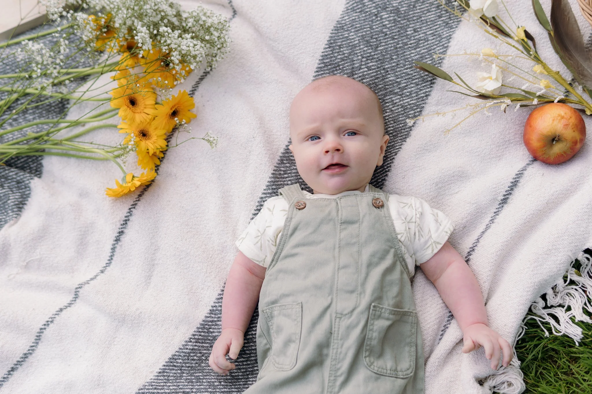Baby ligt op een beige en grijze deken met bloemen links en rechts een appel en bloemen.