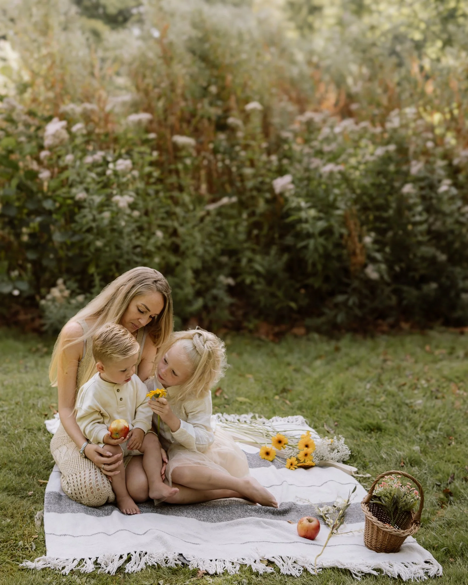 Een vrouw met twee jonge kinderen zit op een witte en grijze gestreepte picknickdeken in een tuin, omgesteld door bloemen en fruit.
