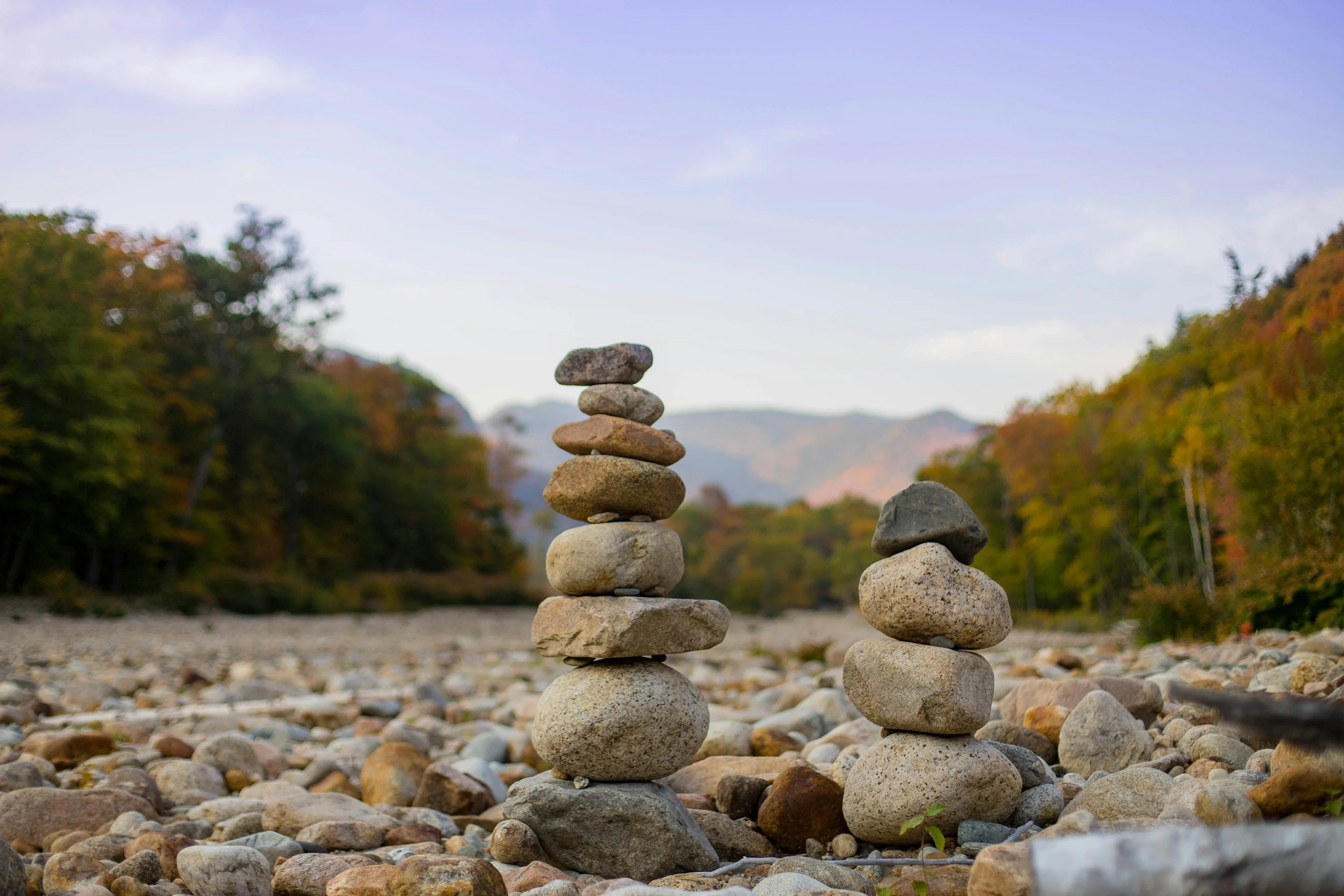 Two stacks of balanced rocks on a rocky riverbank with trees and hills in the background under a partly cloudy sky.