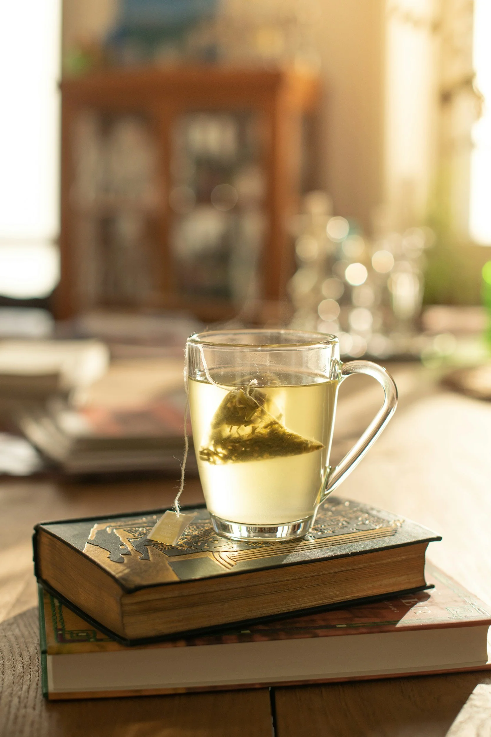 A clear glass mug with a handle contains a tea bag steeping in hot water, resting on a closed book on a wooden table. In the background, there is a blurred bookshelf.