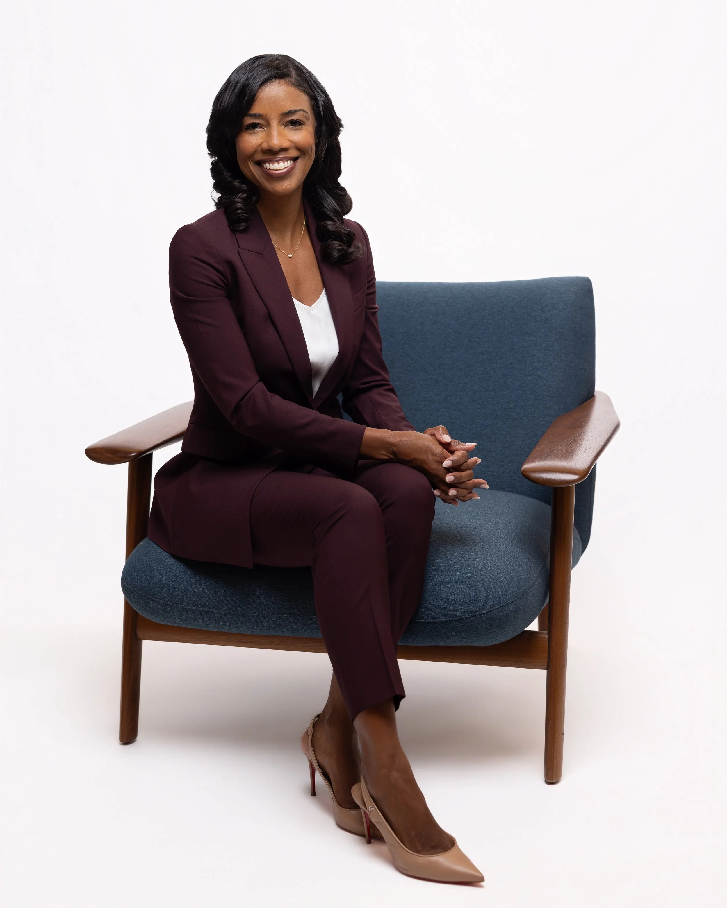A woman with dark hair sitting on a blue chair, smiling, wearing a burgundy suit and beige high heels, against a plain white background.