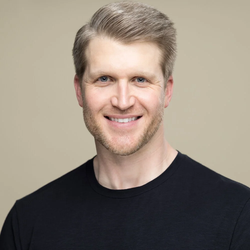 Smiling man with blonde hair wearing a black shirt against a beige background.