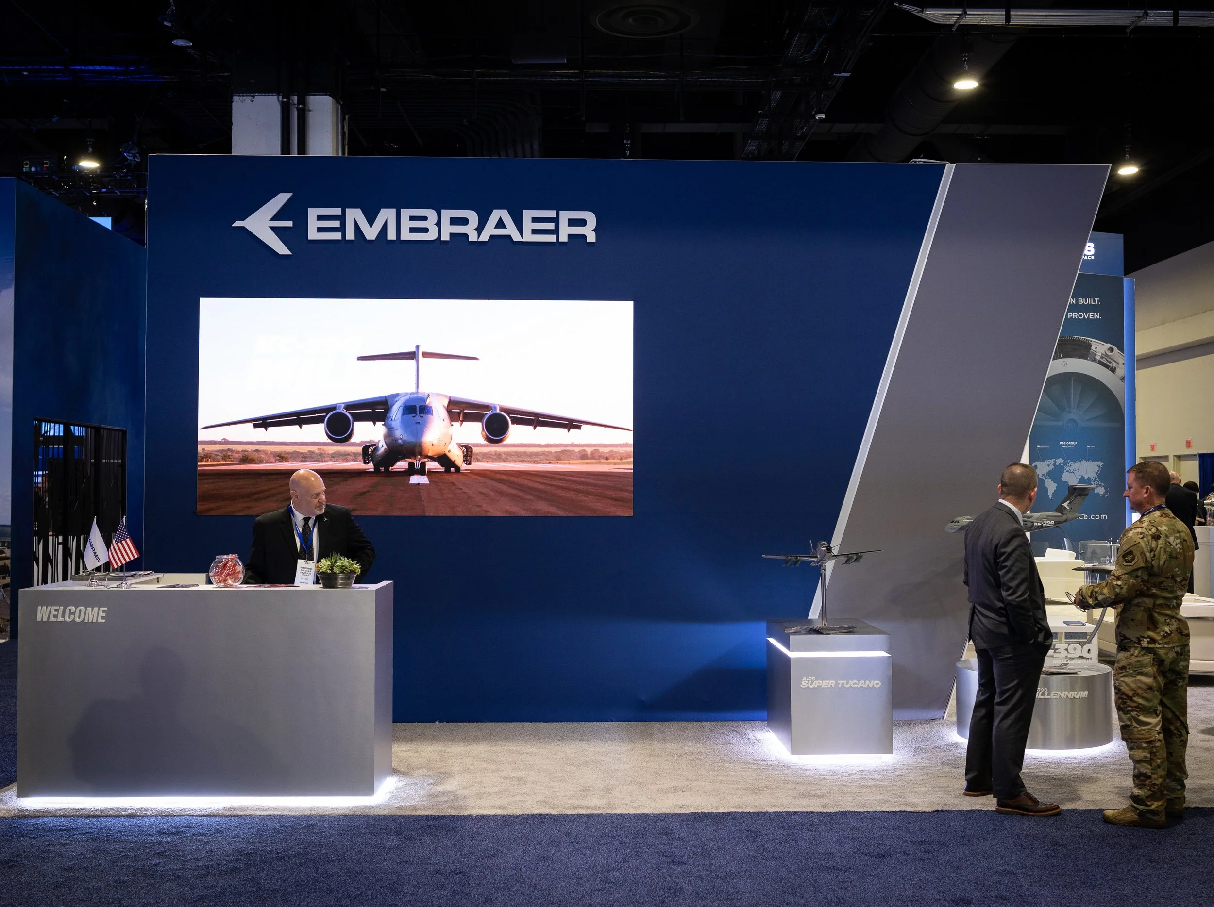 Trade show booth for Embraer featuring a large monitor displaying an aircraft on runway, with people and military personnel engaging nearby.
