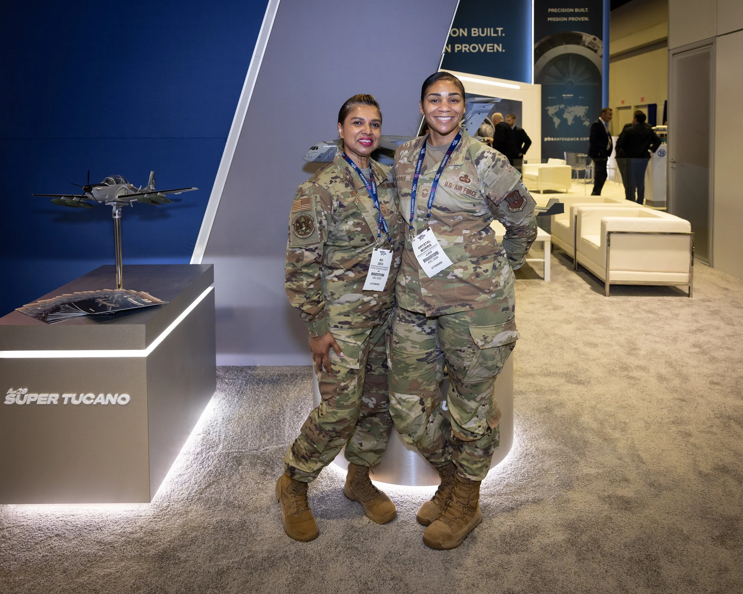 Two women in U.S. Air Force camouflage uniforms smiling at an indoor event, standing in front of a display featuring a model airplane and promotional materials.