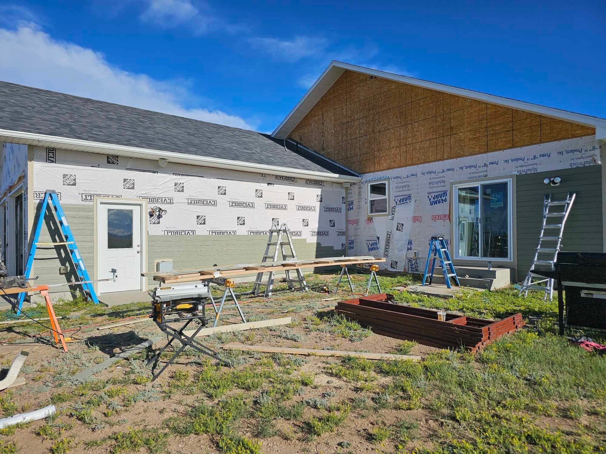 A house under construction with siding partially installed, ladders, construction tools, and building materials in the yard, with a blue sky overhead.