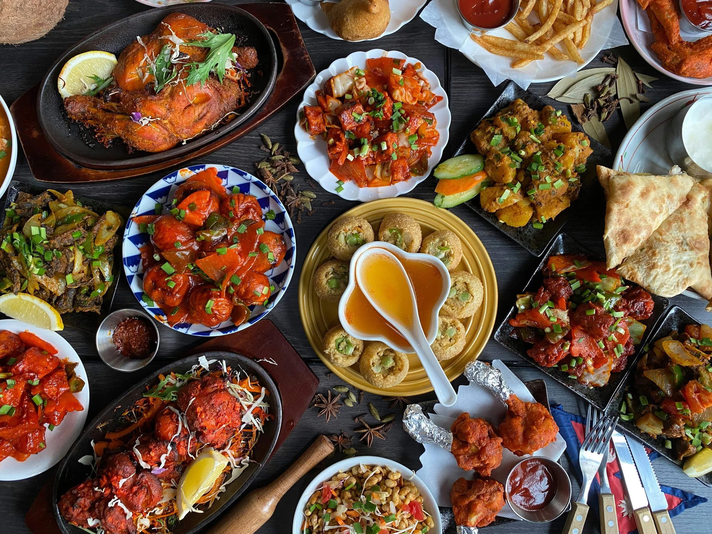Assorted Indian dishes on a table, including chicken curry, vegetable biryani, paneer, samosas, papadum, and fried chicken, with condiments and garnishes.