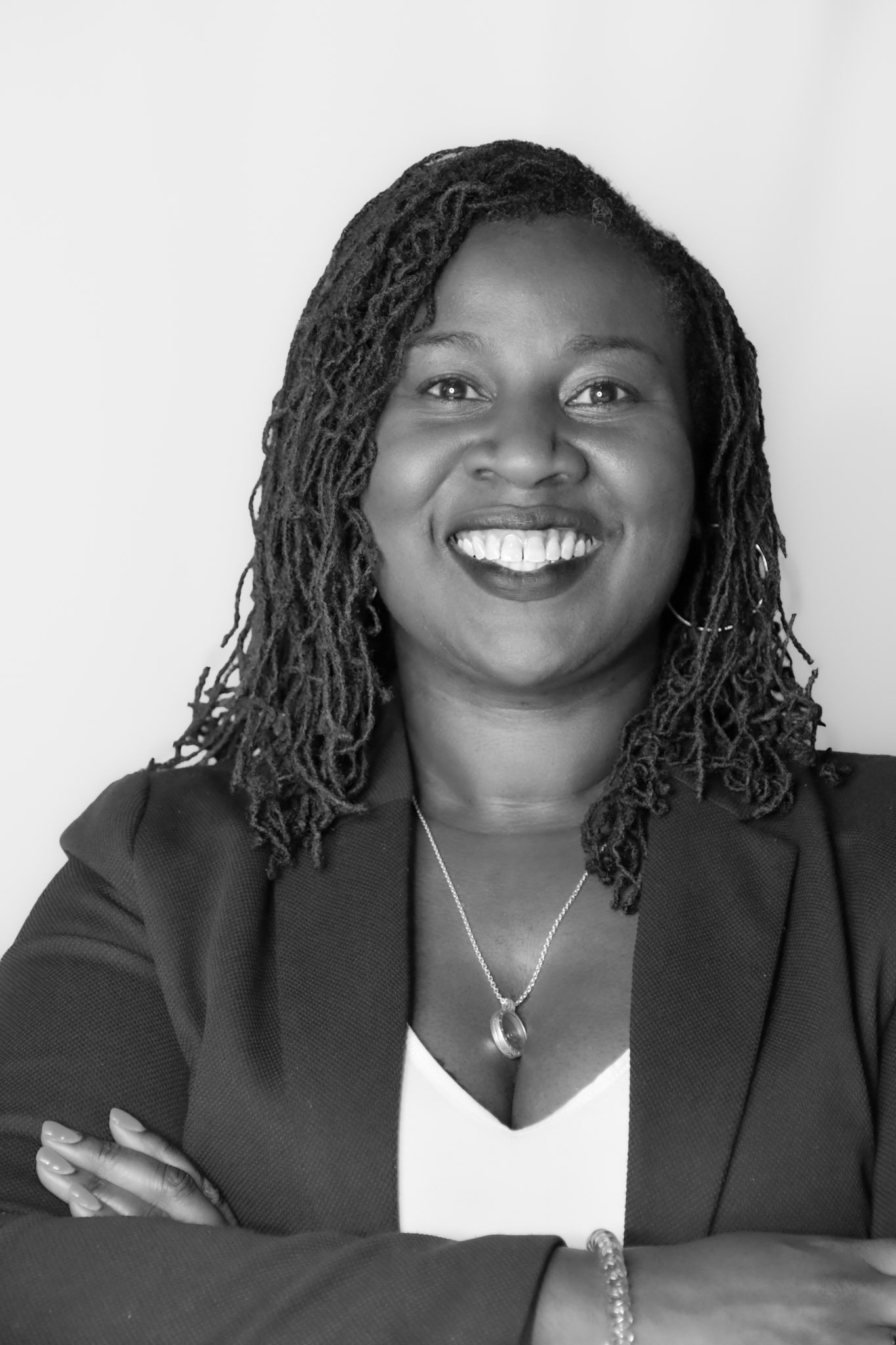 Black and white professional portrait of a woman with shoulder-length dreadlocks, smiling, wearing a blazer, a necklace, and a bracelet, with arms crossed.