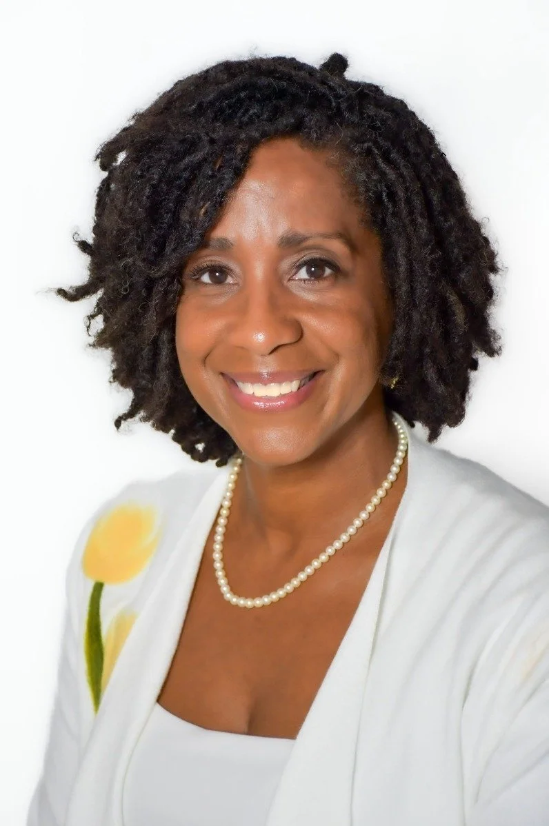 A smiling woman with natural curly black hair wearing a white top, a pearl necklace, and a white jacket with a yellow flower on the shoulder, against a white background.