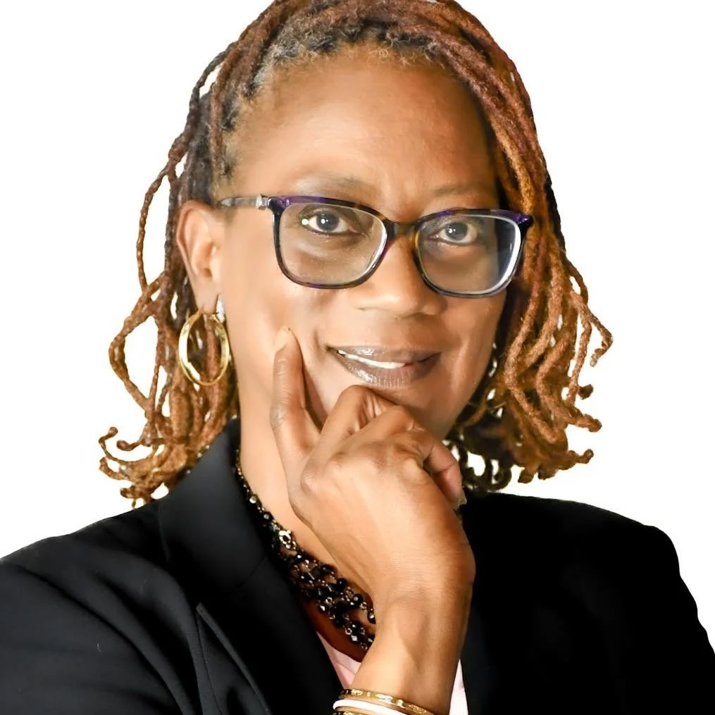 A woman with shoulder-length dreadlocks, wearing glasses, gold hoop earrings, and a black blazer, resting her chin on her hand and looking at the camera with a smile.