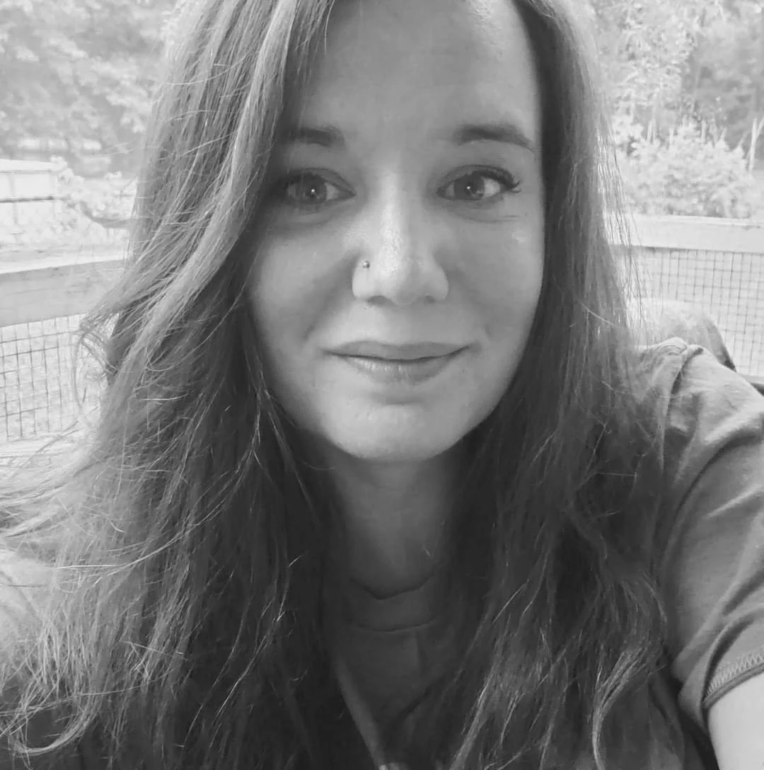 A woman with long hair and a nose piercing smiling at the camera, sitting outdoors with a fence and trees in the background.