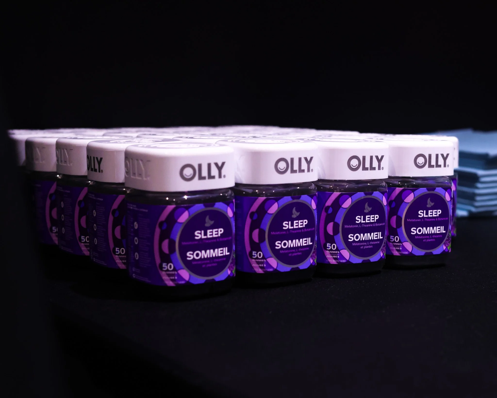 Rows of OLLY Sleep gummy supplement jars with purple and black labels and white lids, stacked neatly on a black surface under bright lighting.