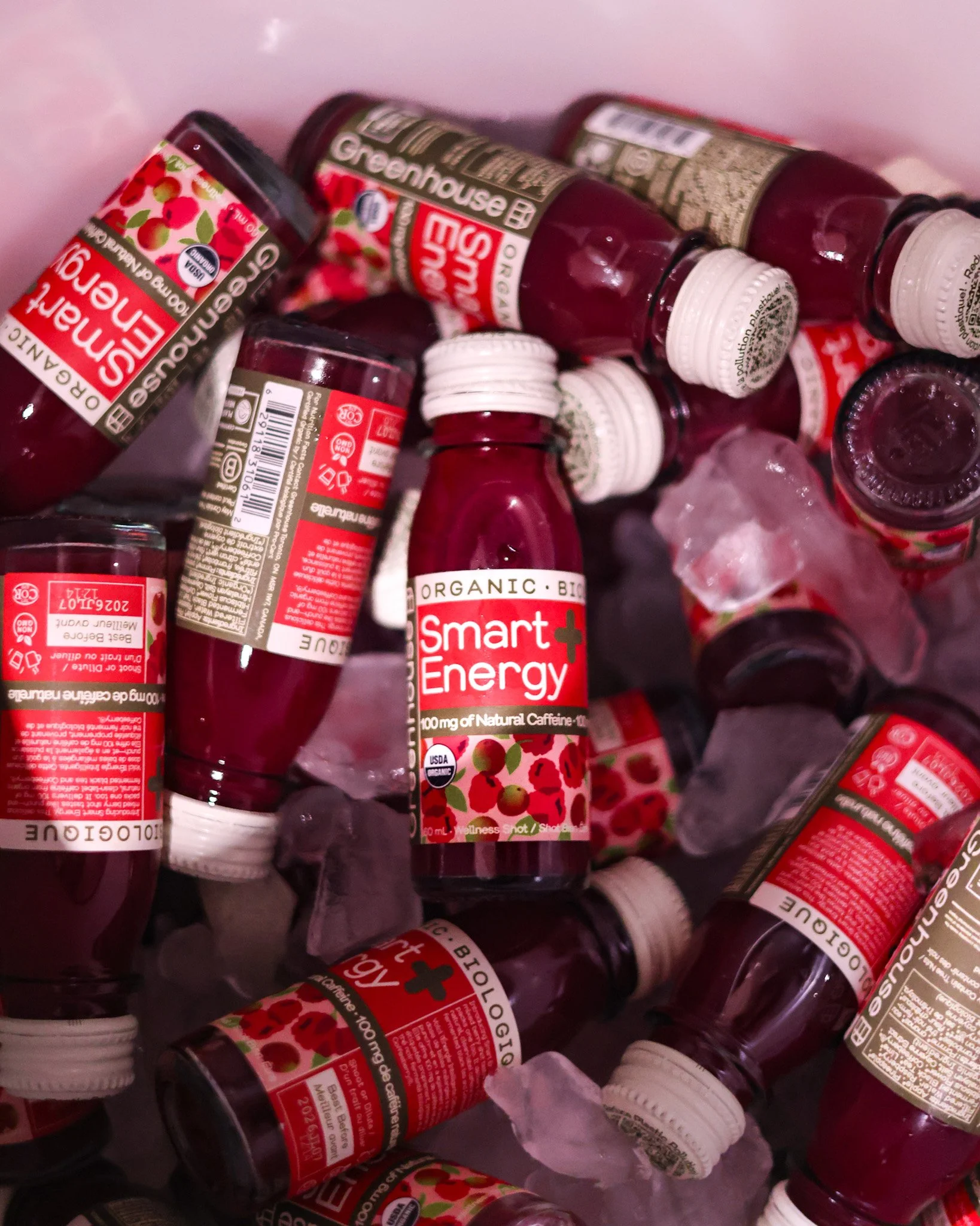 Close-up of multiple bottles of Greenhouse Organic Smart Energy shots packed in ice, with red and white labels featuring cherry illustrations and text highlighting 100mg of natural caffeine.