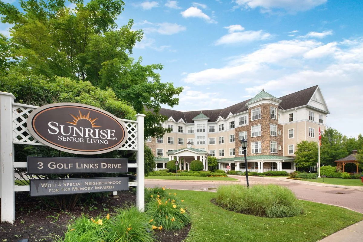 Exterior view of Sunrise Senior Living at 3 Golf Links Drive, with a large sign in the foreground and a beautifully maintained building and garden behind it. This location hosts pudgy pups puppy therapy sessions for senior residents.