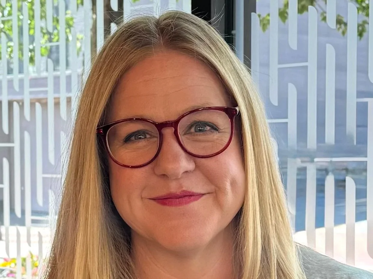 A woman with long blonde hair wearing red glasses, smiling indoors near a window with geometric patterns and green trees outside.