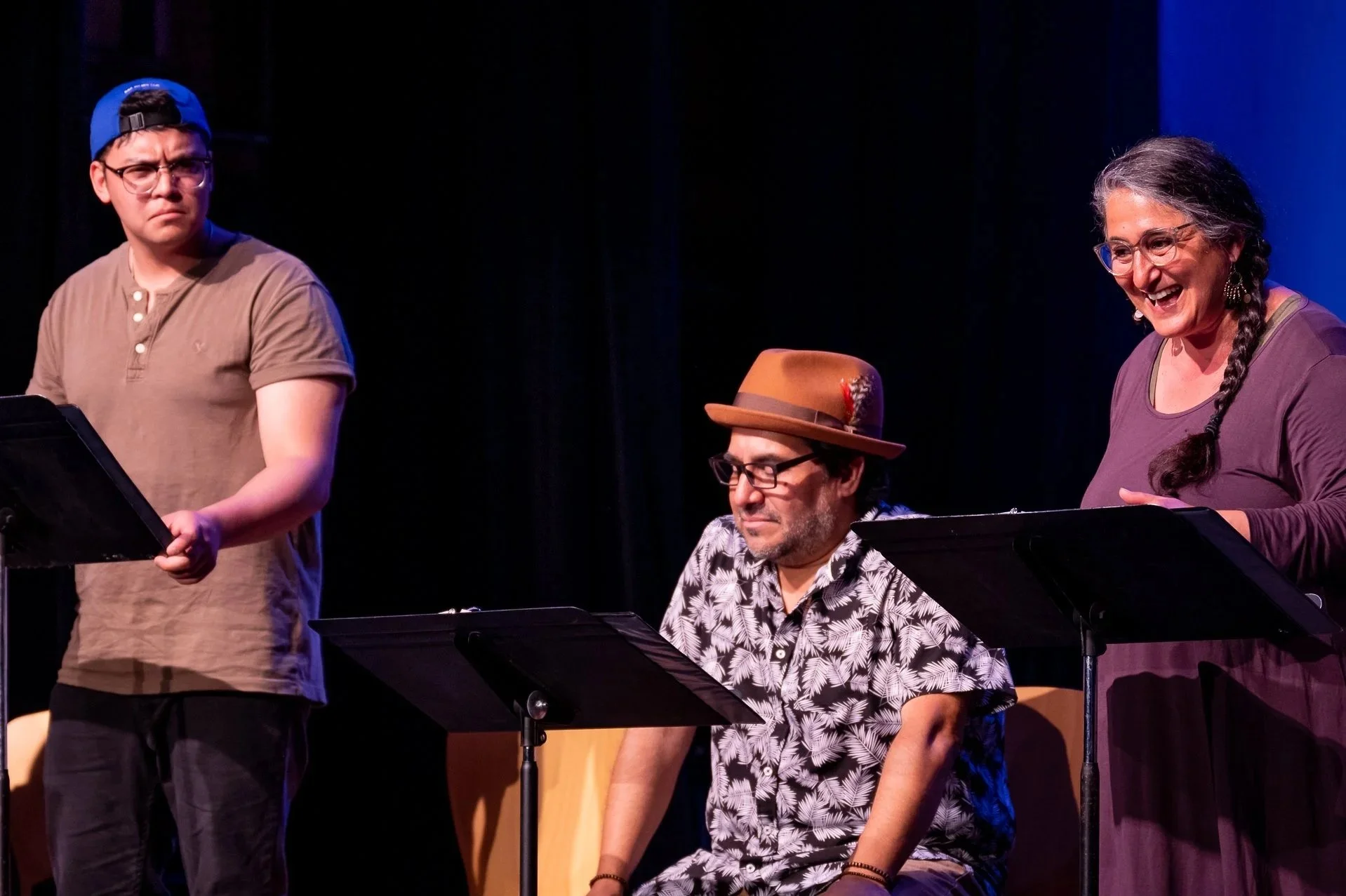 Three people on stage during a reading or performance, each standing or sitting in front of a music stand with scripts. One person is wearing a backwards blue cap, another has a brown hat with a feather, and the third has long hair braided and is smi