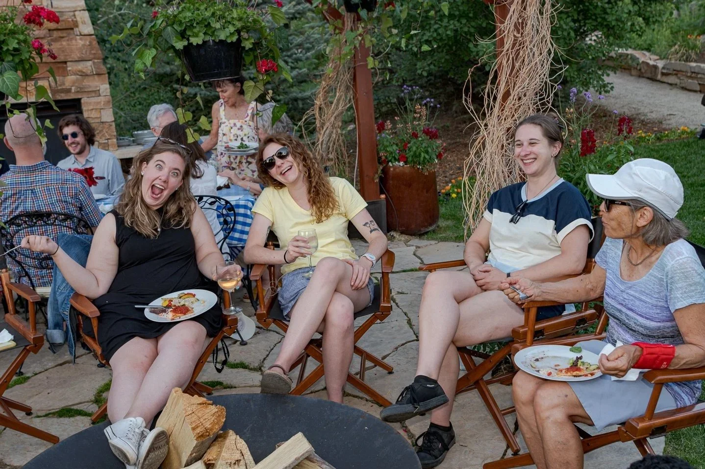 Group of people sitting around a fire pit outdoors, smiling and enjoying a social gathering with food and drinks. Hanging plants and garden visible in background.