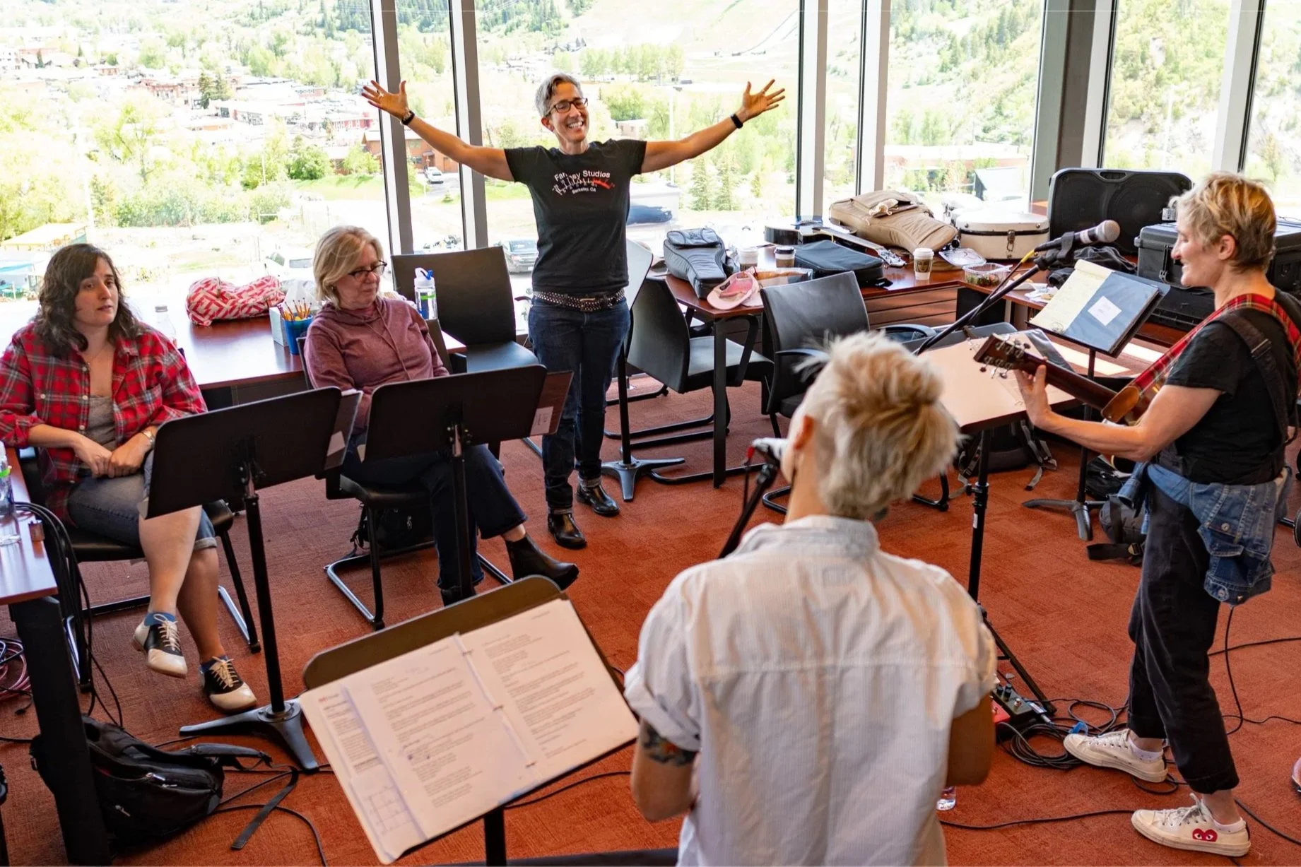 A group of people in a music rehearsal room, with two individuals standing and singing while playing guitar, and others sitting with music stands and notes, in front of large windows overlooking a scenic landscape.
