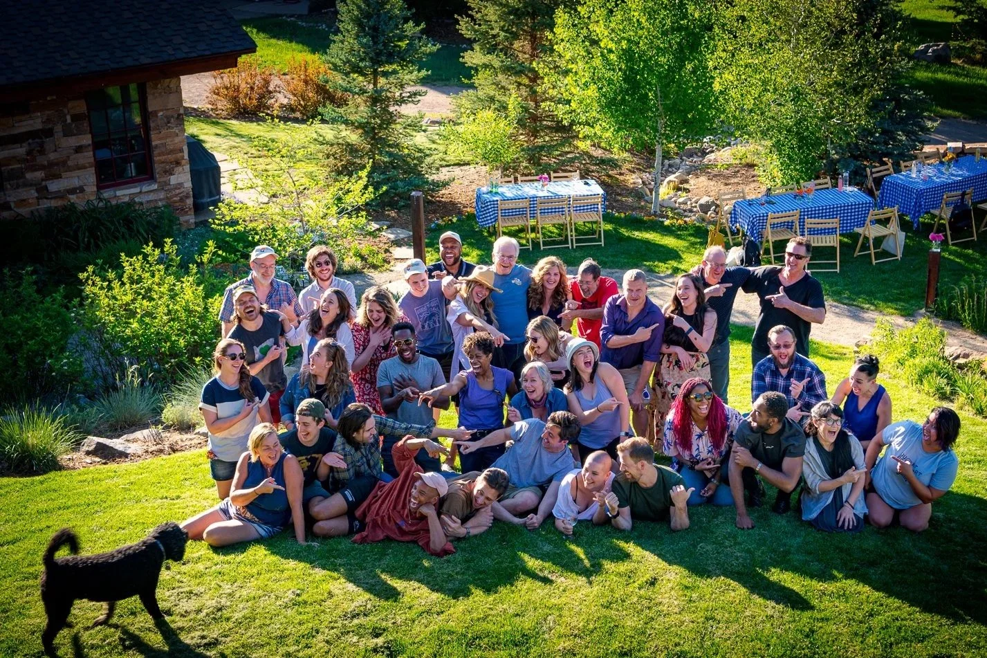 Group of people with playful poses on grassy area, tables with checkered cloths in background, trees and shrubs surrounding them, and a black dog nearby.